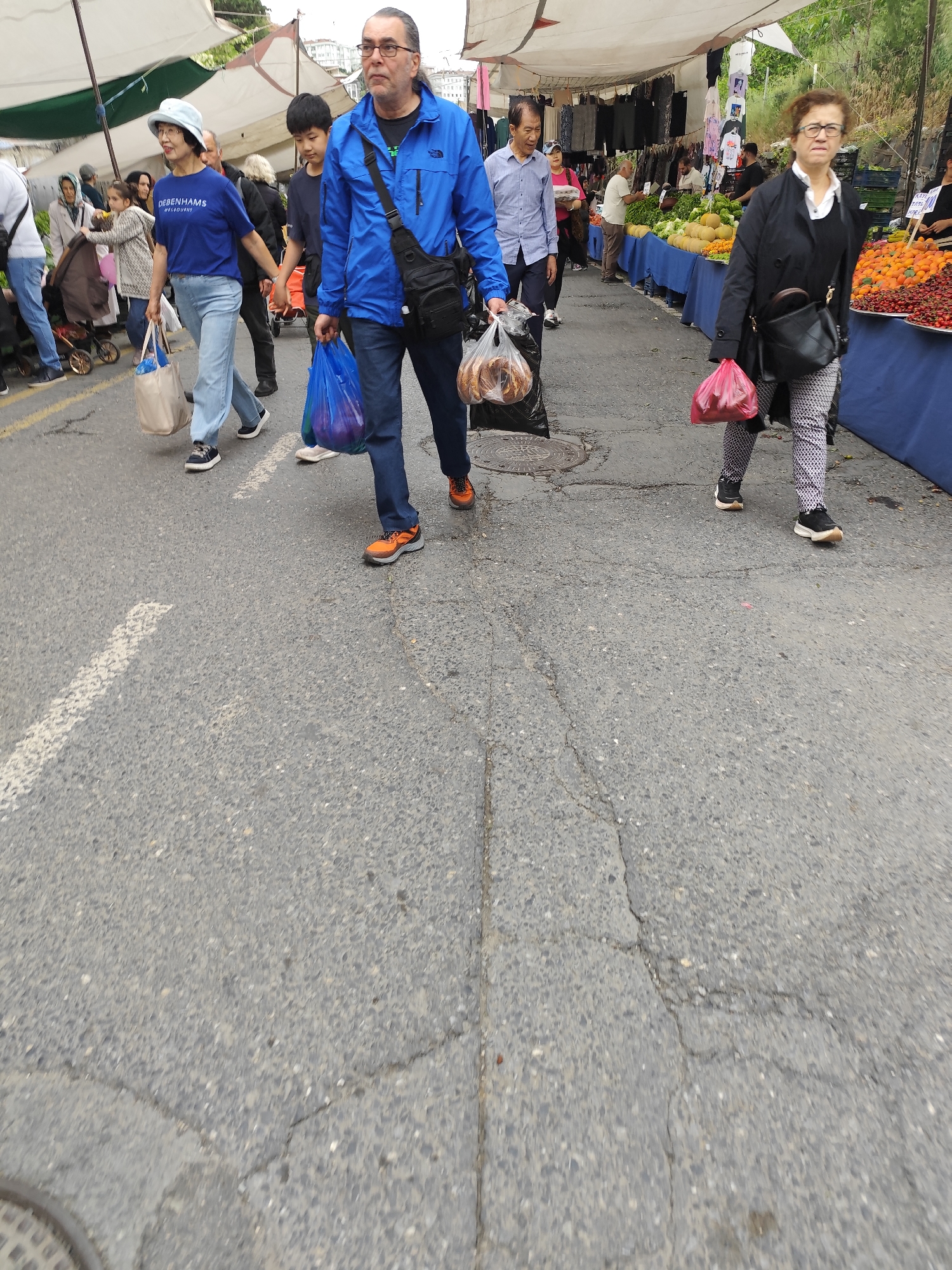 The image depicts a street market scene in an unidentified city. Several people are walking along the street, carrying bags of groceries. Market stalls line the sides of the street, displaying fruits, vegetables, and clothing. The street is paved and shows signs of wear, with cracks and patches visible. The atmosphere suggests a bustling local market.