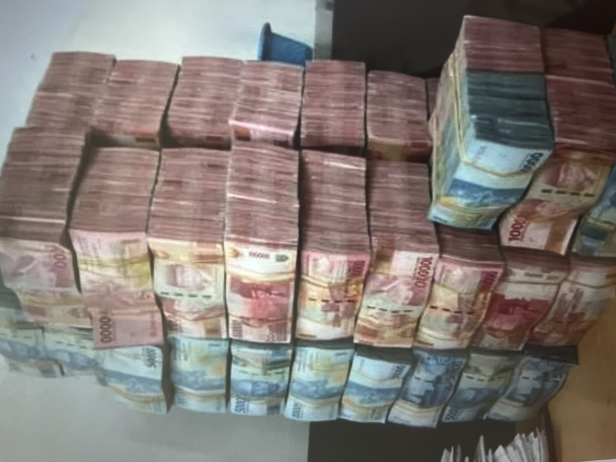 The image shows a large collection of stacked banknotes, likely currency. The banknotes are predominantly pink and red, with some stacks also featuring green and blue hues. Several stacks have visible text, including "10000", "100", and "5000". The money appears to be neatly stacked and organized in rows, suggesting it might be in a storage or display setting. The lighting suggests an indoor environment, possibly a room or a space where the money is being kept. There are no people visible in the image, and the focus is solely on the stacks of money. The arrangement and sheer volume of the currency imply a significant amount of money. The image is slightly blurry, making it difficult to discern finer details of the banknotes or the surrounding environment.