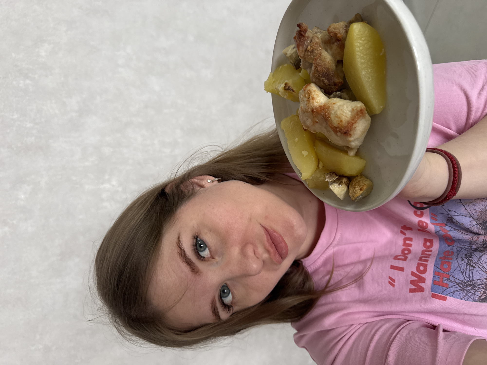 A female individual is depicted in a medium shot, holding a plate of food. She has long brown hair, blue eyes, and fair skin, with a slight pout on her lips, looking directly at the camera. She wears a pink short-sleeved t-shirt with red text that partially reads, "I Don't Wanna Be c" and "I Hate..." above a graphic of bare trees. A small silver hoop earring is visible in her left ear, and a red elastic band is on her right wrist. She holds a white, circular plate containing several pieces of cooked poultry, numerous yellow chunks of boiled or roasted potatoes, and smaller, darker, irregular pieces that resemble mushrooms. The background consists of a plain, light grey or off-white wall exhibiting a subtle, mottled texture. The scene is located in Gornyi (Dvurechenskoe S/P), Russia.