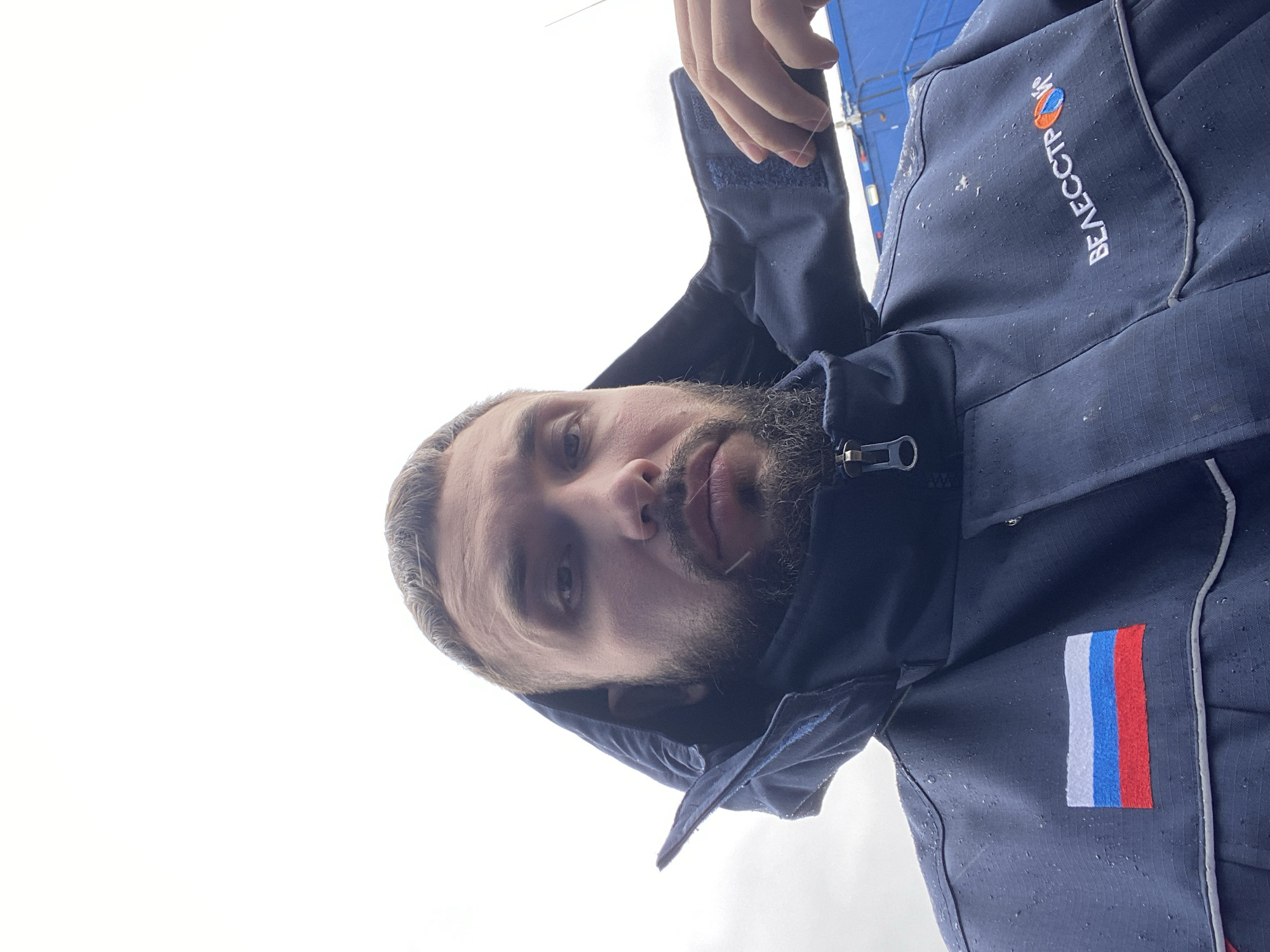 A close-up, tilted selfie shows a man with fair skin, a salt-and-pepper beard, and a graying hairline. He's wearing a dark blue jacket with the name "BEAAECTPCW" embroidered vertically in white on the upper right chest. The jacket has a zipper partially visible at the neckline and what appears to be a hood pulled up around his head. A patch of the Russian flag is visible on the jacket's left sleeve. The background is a bright, overcast sky. There's insufficient information to confirm the image was taken in Luzhitsy, Russia.