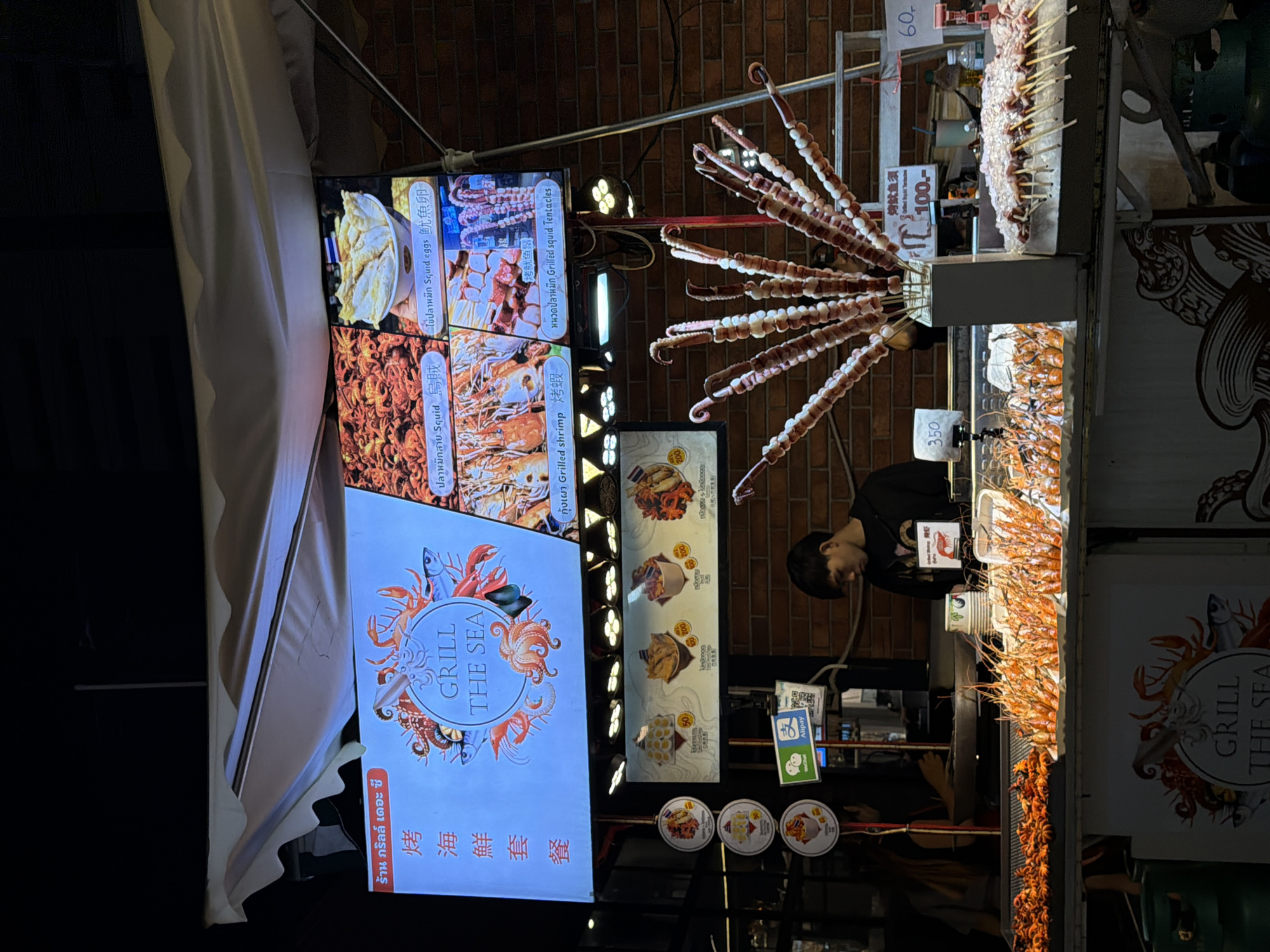 This is an outdoor night market scene in Bangkok, Thailand, likely a food stall focused on seafood. A large digital menu board displays various grilled seafood items with Thai and Chinese text, including images of shrimp, squid, and other sea creatures. Below the menu board, a display case showcases an abundance of grilled shrimp and smaller, bright orange seafood items, possibly baby octopus. To the right, skewers of raw meat are displayed on ice, awaiting grilling. A young man, presumably a vendor, is visible behind the counter, looking down. Several payment apps, including WeChat and Alipay, are advertised on the stall. The overall ambiance suggests a bustling street food environment.