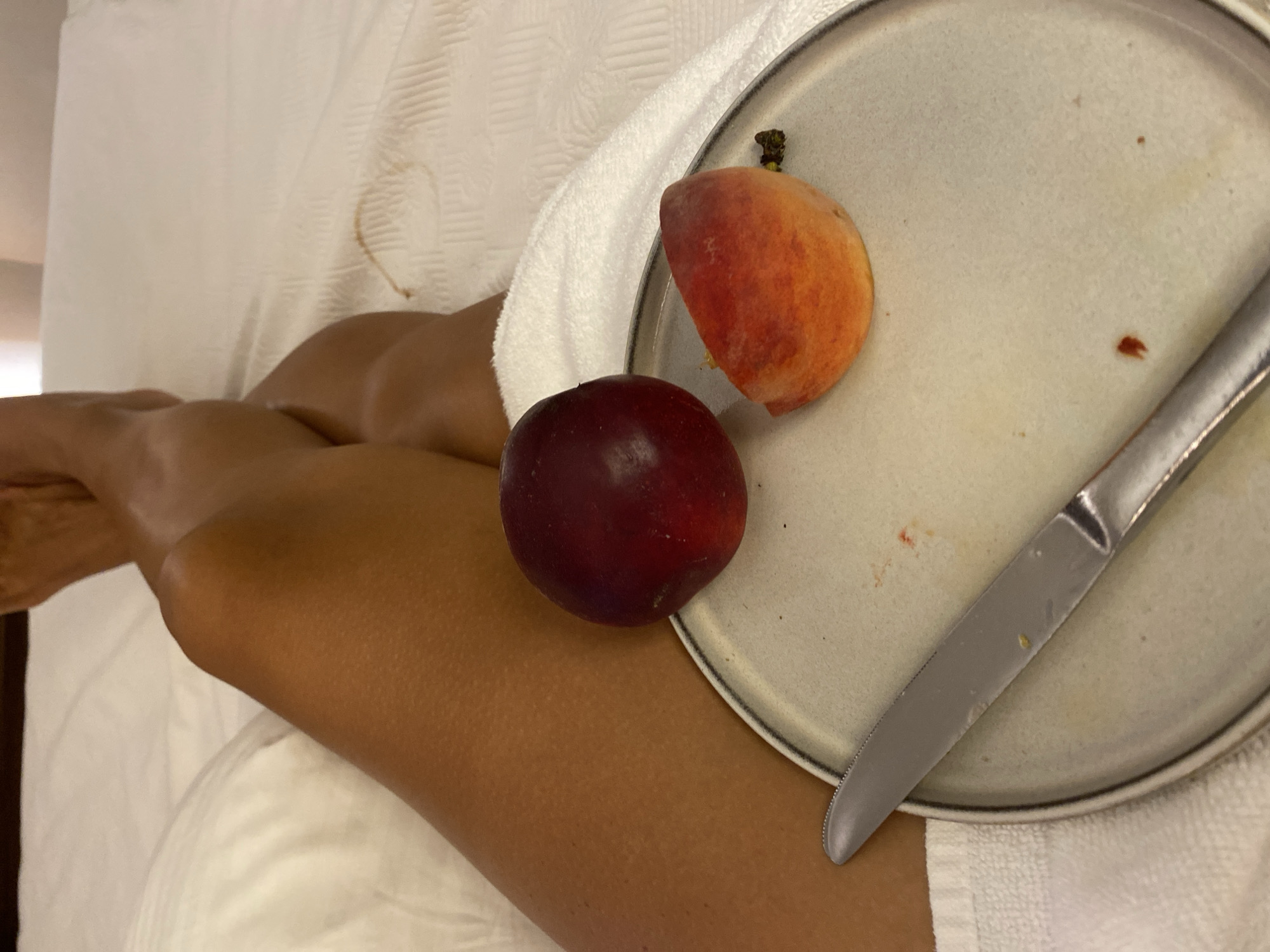 The image shows a person's legs, crossed, lying on a white bed with a white towel draped over them. On a light gray plate in front of the legs are a whole red plum and a slice of peach. A silver knife is placed on the plate. The background shows white bedding and a hint of a dark headboard. There is a brown stain on the white bedding to the right of the legs. The image is taken from a high angle, looking down. There is no indication of the city of Aksu, Turkey.