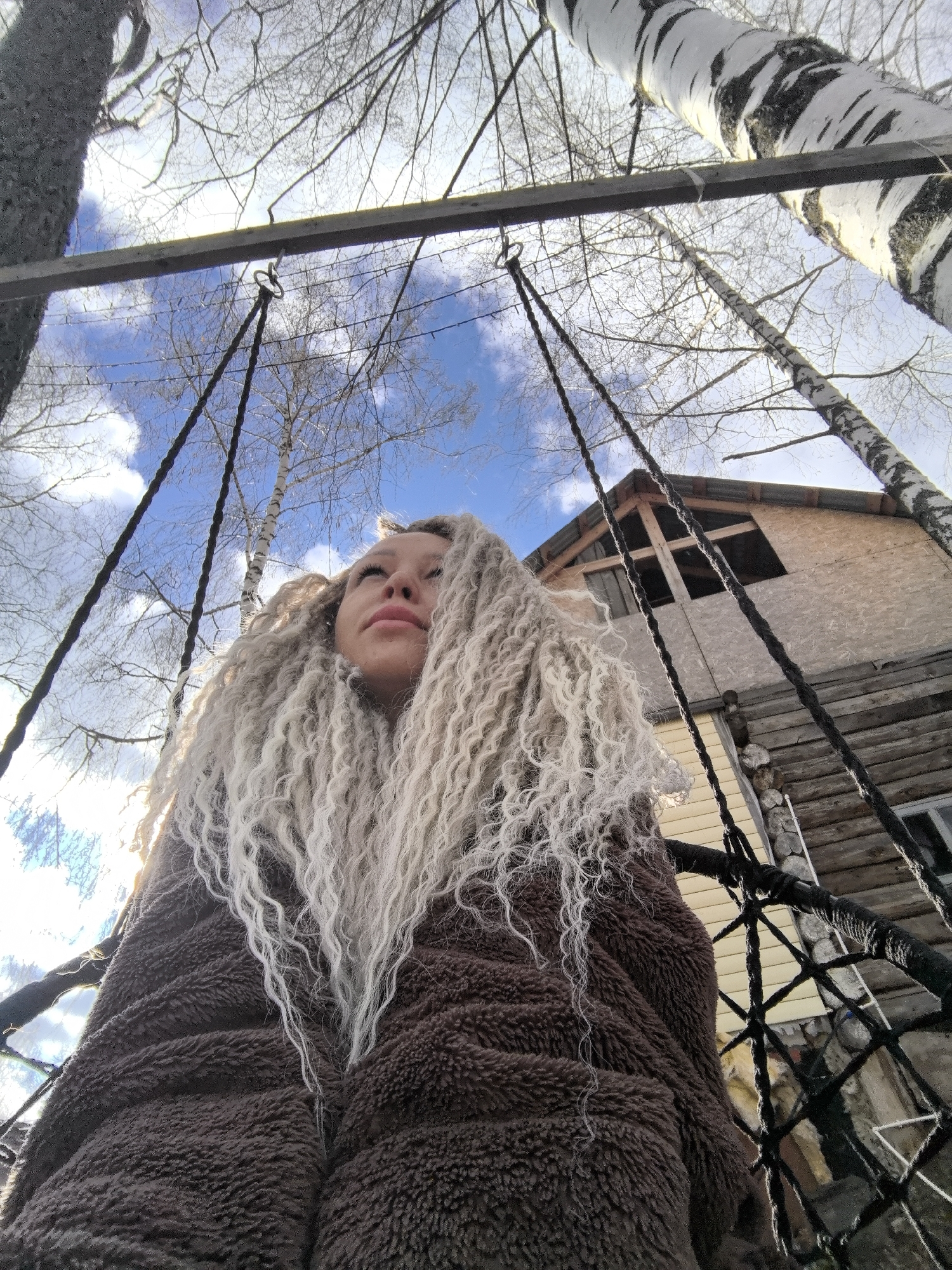 An outdoor, low-angle shot captures a young woman looking upwards, framed by trees and a clear sky. The central subject is a woman with long, voluminous, light blonde or white curly hair, possibly dreadlocks, cascading over her shoulders. She is wearing a thick, textured, brown fuzzy jacket or blanket. Her gaze is directed towards the bright sky, suggesting a moment of contemplation or relaxation.
She appears to be on a swing, with dark, thick ropes visible extending upwards from either side of her towards a horizontal wooden beam overhead. A metal ring secures one rope to the beam. The background features a vibrant blue sky with scattered white clouds. Bare deciduous trees, including several distinctive white and black-barked birches, punctuate the upper and left parts of the frame, their branches reaching towards the sky.
To the right, partially visible behind the swing ropes, is a two-story wooden house. The upper section appears to be constructed from light-colored panels with a prominent cross-shaped window frame, while the lower section features log construction. The scene suggests a daytime setting, likely late winter or early spring given the bare trees, with bright sunlight. The rustic elements, including the birch trees and wooden house, align with the provided location context of Noviy, Russia. No visible text is present in the image.