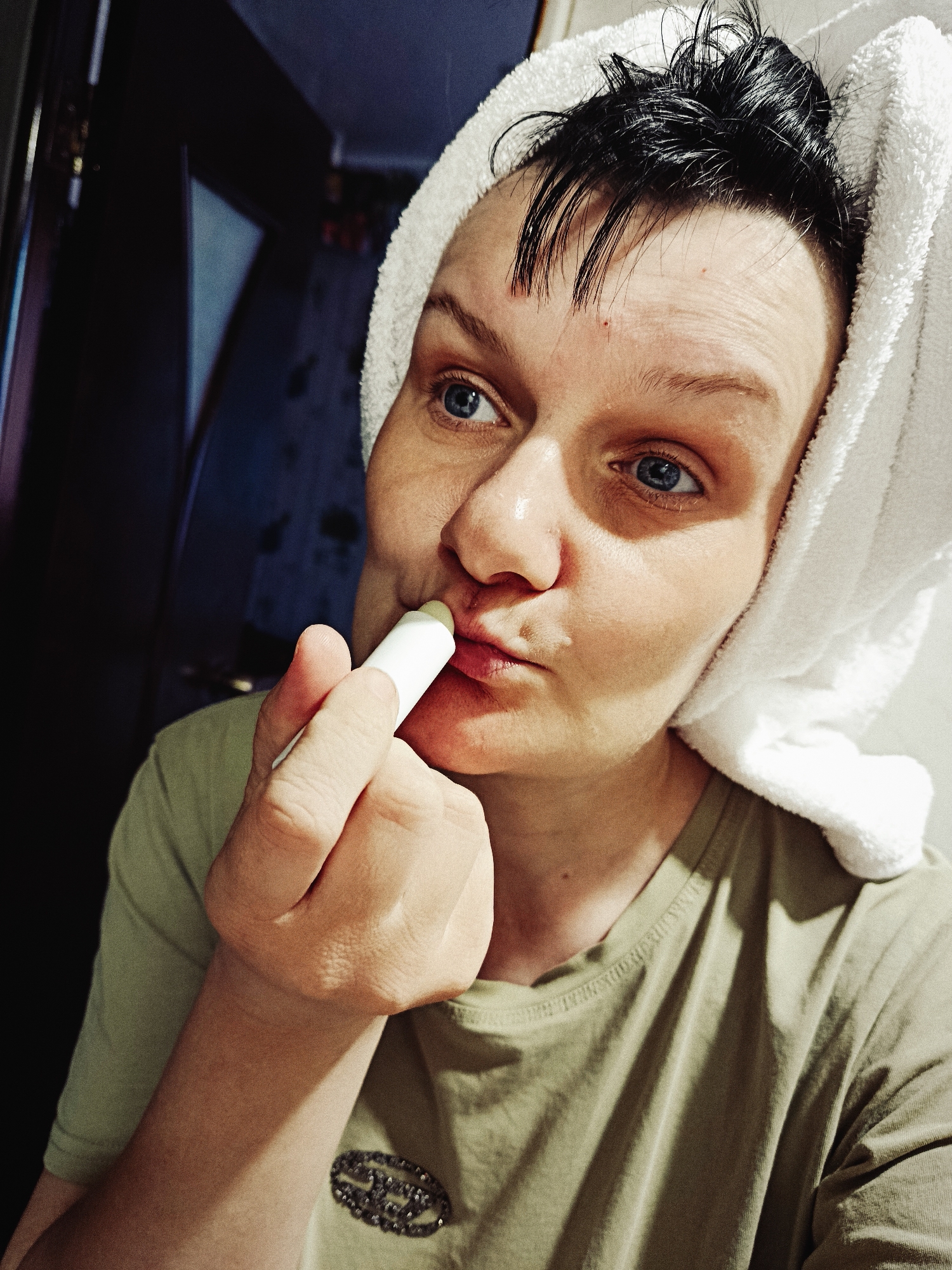 A woman with wet dark hair partly covered by a white towel is applying lip balm. Her eyes are blue, and she's wearing a green t-shirt. The background shows a door and part of a room. The location is not discernible from the image.