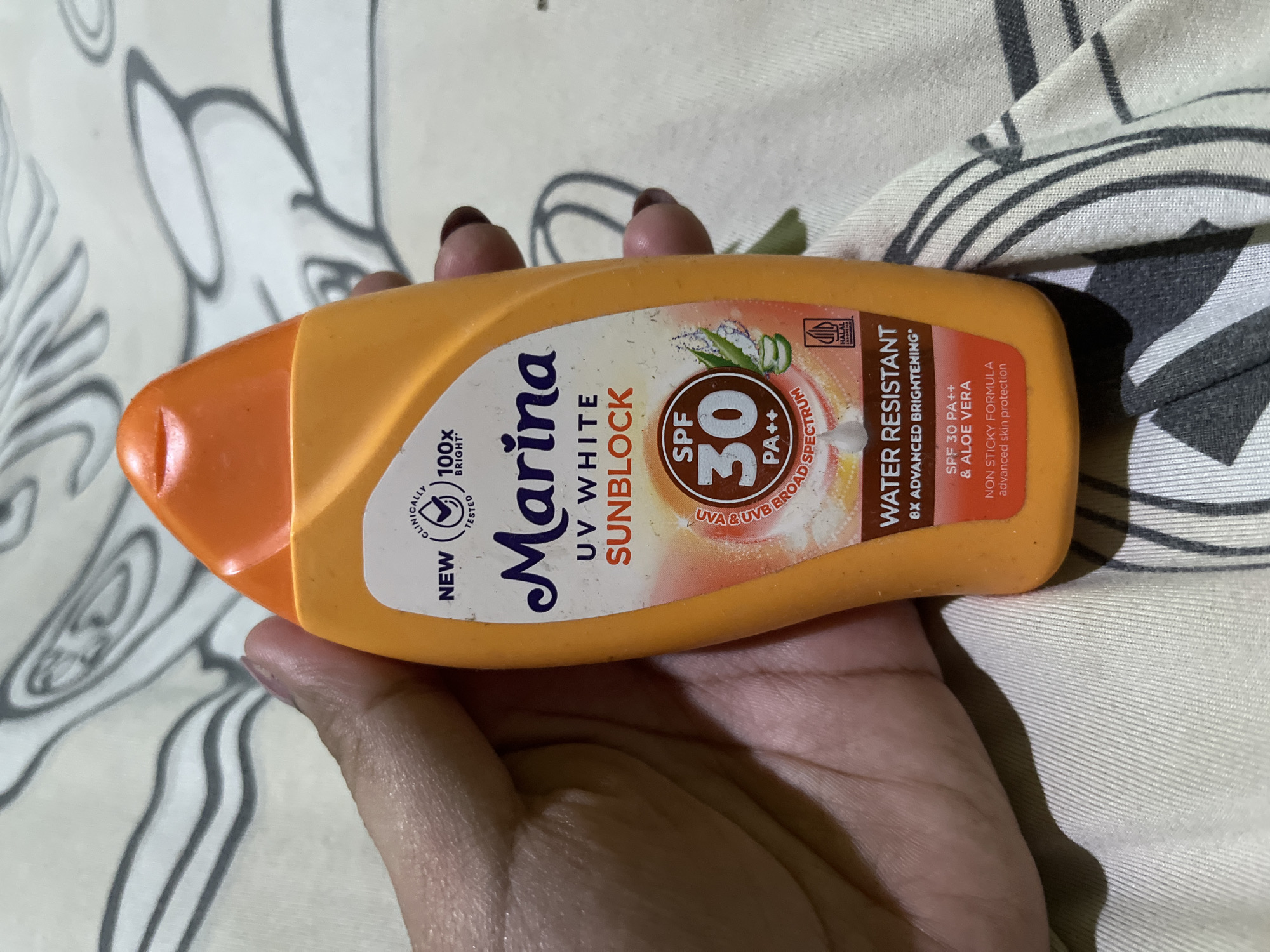 An orange sunscreen bottle with an orange cap is held in a person's left hand against a light-colored fabric background with a grey abstract pattern. The bottle has a white label with blue and orange text. Prominently displayed on the label is the brand name "Marina" in a cursive font, followed by "UV WHITE SUNBLOCK". Below this, there is a circular emblem with "SPF 30 PA++" in the center, surrounded by graphics of water drops, aloe vera, and shimmering light effects. Other text on the label includes "NEW", "CLINICALLY TESTED", "100x BRIGHT", "WATER RESISTANT", "8X ADVANCED BRIGHTENING", "SPF 30 PA++ & ALOE VERA", and "NON STICKY FORMULA". A small square logo with "HALAL INDONESIA" is visible at the bottom right of the label. The background fabric appears to be a patterned bedsheet or similar textile. The image was taken at an eye-level perspective. There is no information about the location in Bumi Mulyo, Indonesia.