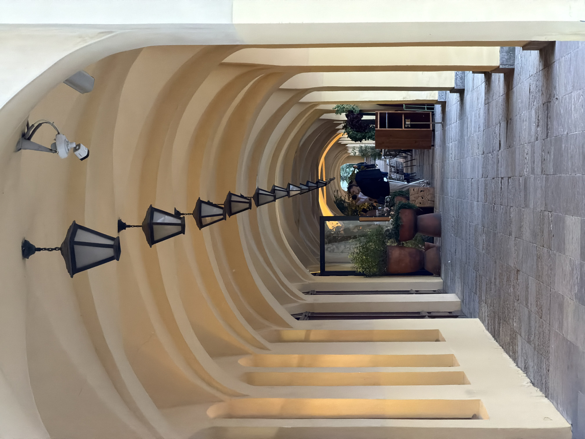 The image depicts a long, arched colonnade with a stone-tiled floor. Several black, hexagonal lanterns hang from the arched ceiling, spaced at intervals down the walkway. A white security camera is mounted on the ceiling near the entrance of the colonnade. The walls of the colonnade are painted a light yellow. On the left side, there are recessed arched openings. On the right side, further down the colonnade, tables and chairs are visible, suggesting an outdoor seating area for a cafe or restaurant. Several potted plants are placed along the walkway. Two individuals are partially visible in the distance, walking away from the camera. The lighting suggests it is daytime. The style of architecture and the presence of elements like the colonnade and the lanterns are common in historical city centers, potentially aligning with architectural styles found in cities like Saint Petersburg, Russia, known for its neoclassical and baroque structures.