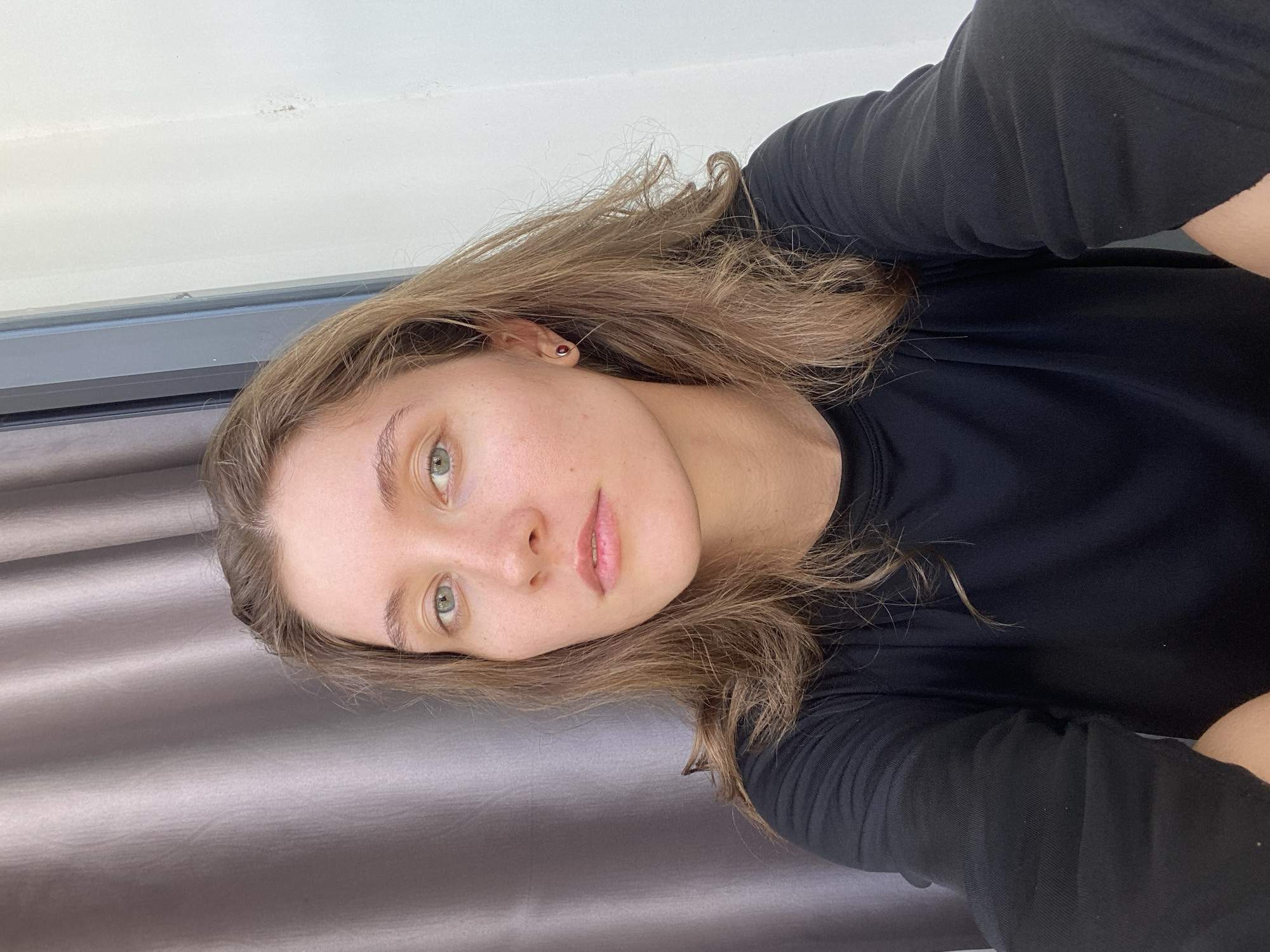 A young woman with long, wavy brown hair and light green eyes is pictured in a close-up, seated selfie. She is wearing a black long-sleeved shirt. A grey curtain is visible in the background to the left, and a white wall with a window frame is on the right. The woman is wearing a small, dark red stud earring in her left ear.