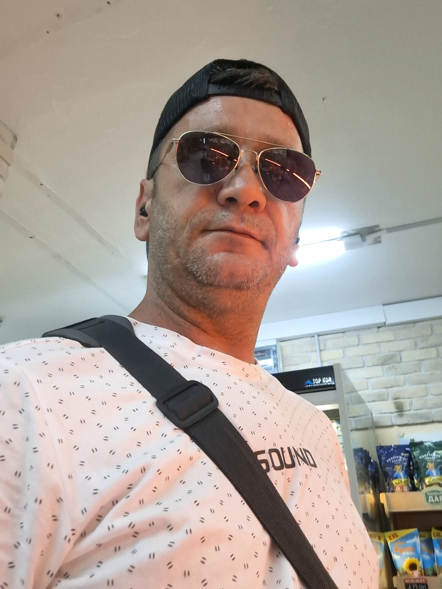 A man wearing sunglasses and a black baseball cap is taking a selfie. He has a light beard and is wearing a white t-shirt with a repeating pattern of double dashes. A dark-colored strap from a bag crosses his chest. In the background, there is a brick wall and shelving units stocked with various products. A sign that reads "TOP KOR" is visible. The lighting suggests an indoor environment, possibly a store. The scene is set in Yerocham, Israel.