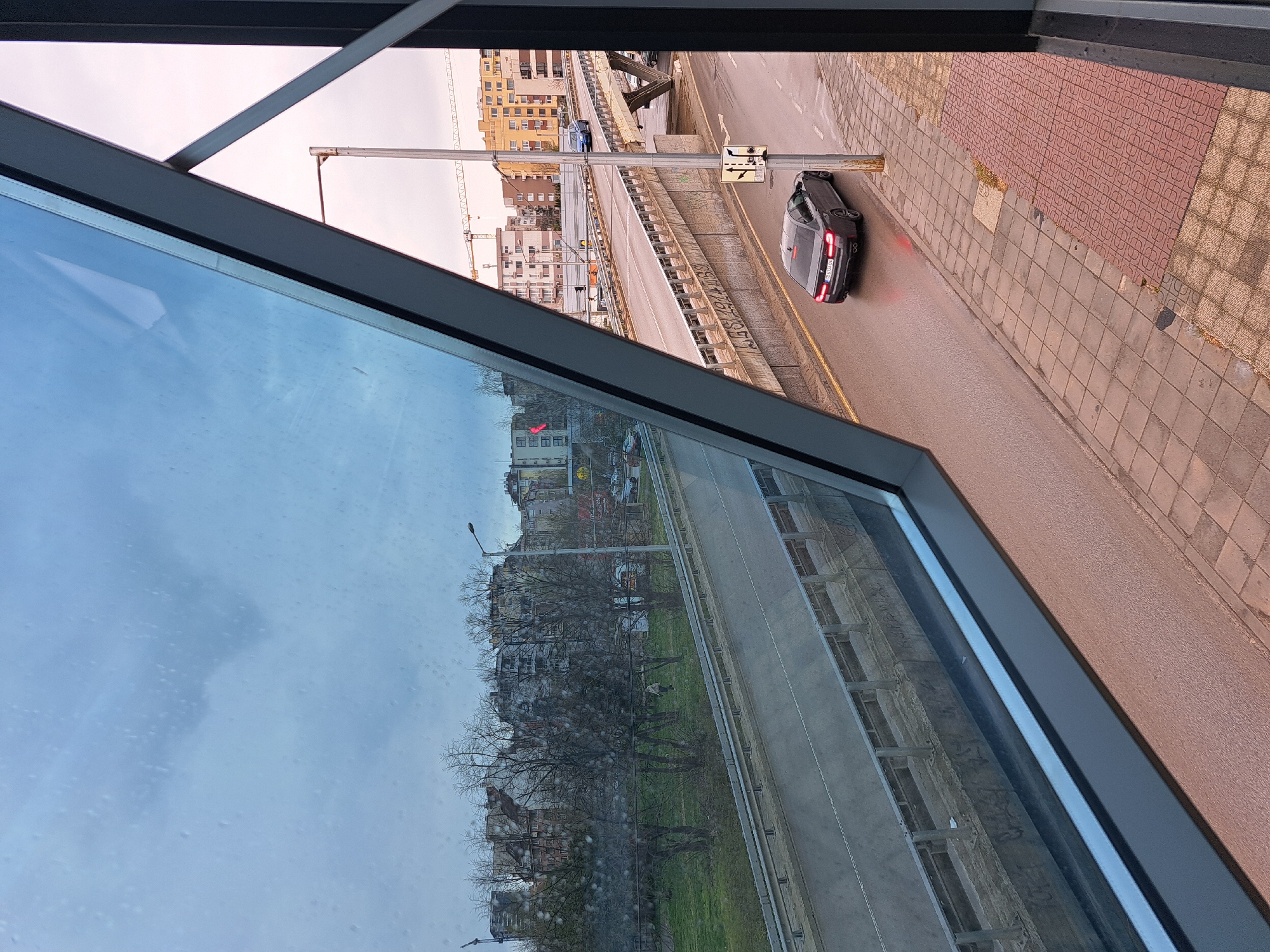 View from a window of a city street with a bridge overhead. A car is driving on the street. The sky is overcast. In the reflection of the window, there are buildings and trees. Location: Unknown, Unknown.