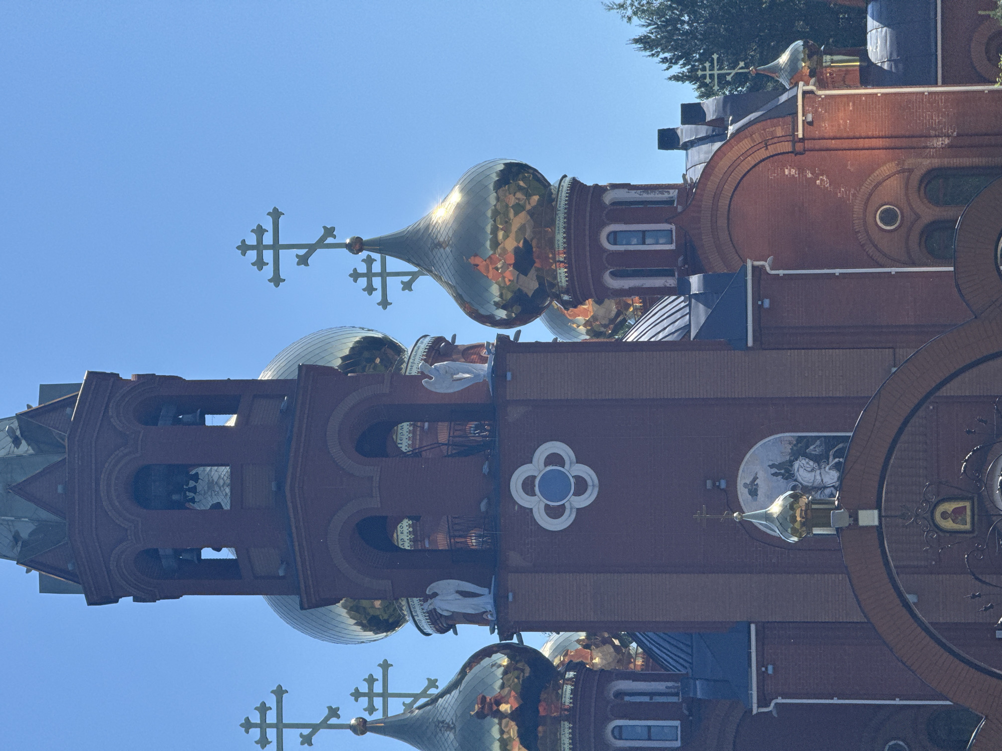 This image shows an exterior view of the Church of the Holy Trinity in Chaikovsky, Russia. The church is constructed primarily of red brick, with several golden domes topped with Orthodox crosses. A prominent bell tower with arched openings and bells is visible on the left side of the image. Decorative architectural elements, including white sculpted angels and a clover-shaped motif, are present on the facade. The sky is clear and blue.