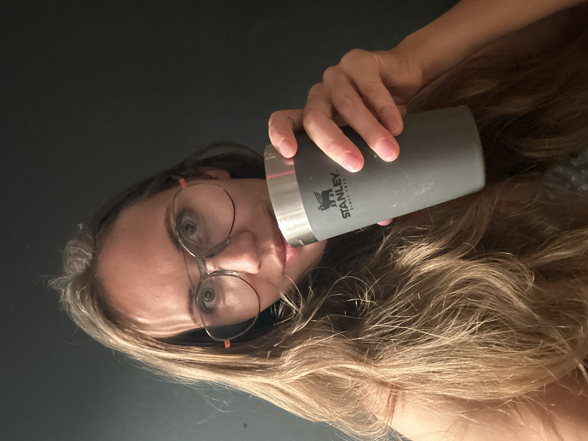 A young woman with long, blonde hair and glasses is holding a grey Stanley brand cup to her lips. She is positioned in the foreground against a dark, plain background. The lighting suggests an indoor setting, possibly a home, with light illuminating her face and hair from the left. There are no discernible landmarks or elements that would indicate a specific location in Fethiye, Turkey.