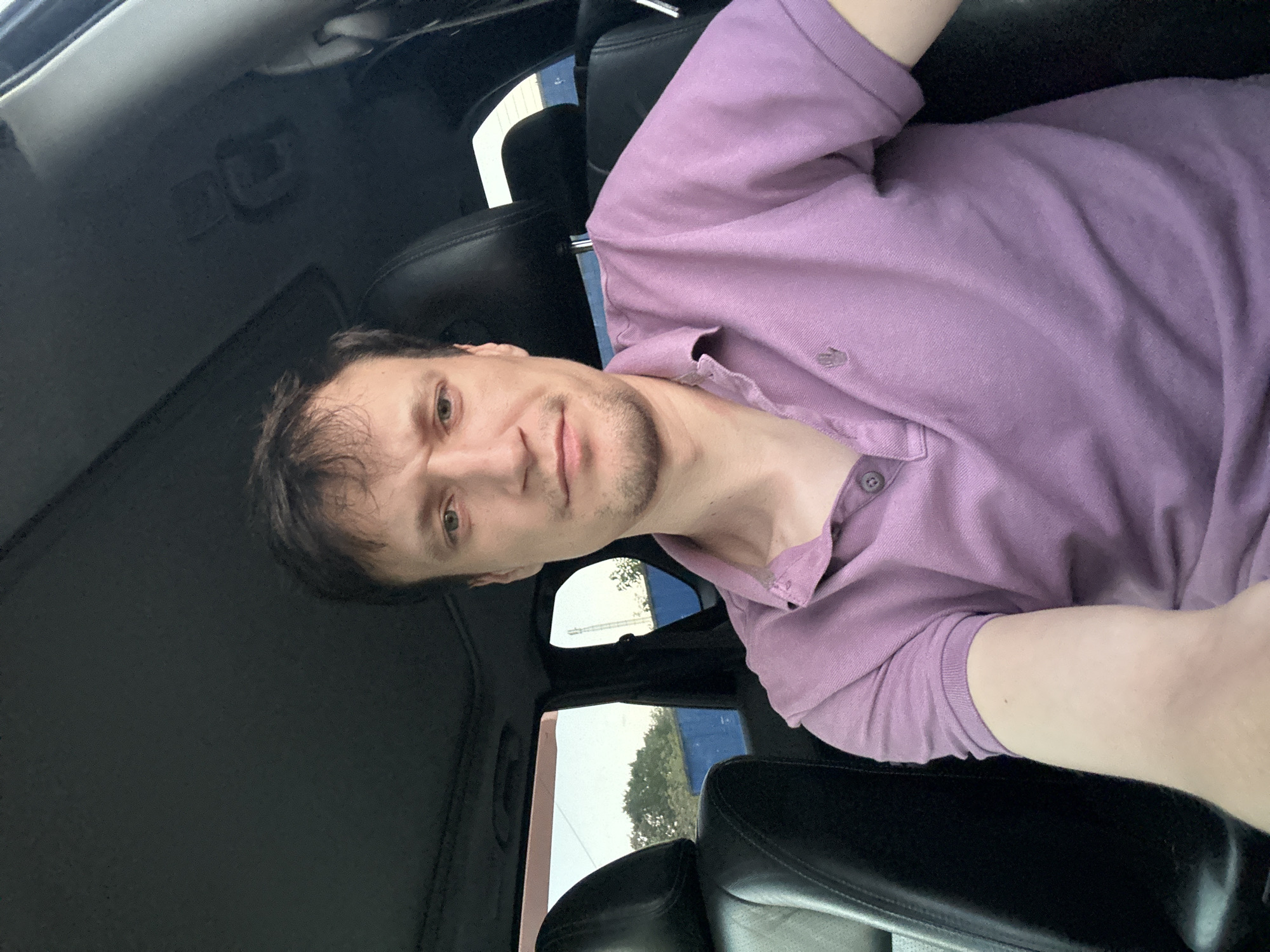 A man with light brown hair and a fair complexion is sitting in the driver's seat of a car, facing the camera. He is wearing a light purple polo shirt with a small embroidered logo on the left breast. His facial expression is neutral, with a slight frown. The interior of the car is visible, including black leather seats and the car's roof lining. Through the windows, trees and a blue structure are visible, suggesting an outdoor setting. The image appears to have been taken during daylight. There is no specific indication that this location is within the city of Shatava, Ukraine.