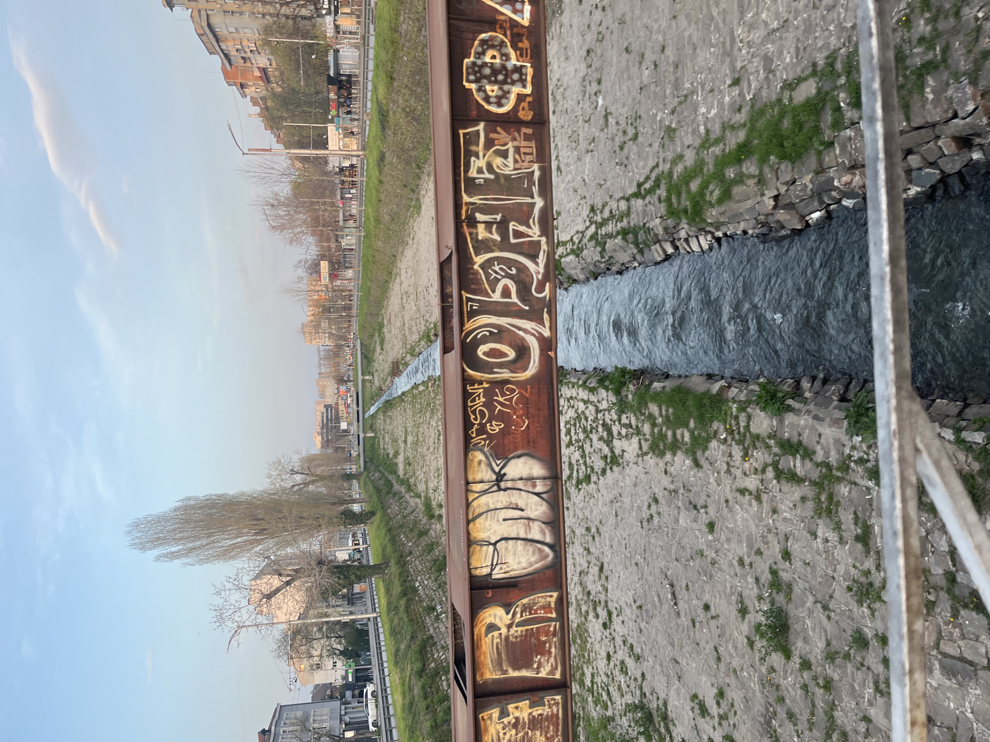 The image is an eye-level shot of Perlovska River as seen from a bridge in Sofia, Bulgaria. The river runs through a narrow concrete channel. The bridge has rusty metal railings covered in graffiti. The banks of the river are mostly concrete and stone with sporadic patches of grass and vegetation. In the background is an urban landscape featuring buildings and trees. The sky is light blue with a few clouds. There are no people visible in the immediate foreground, but there appear to be people walking in the distance.