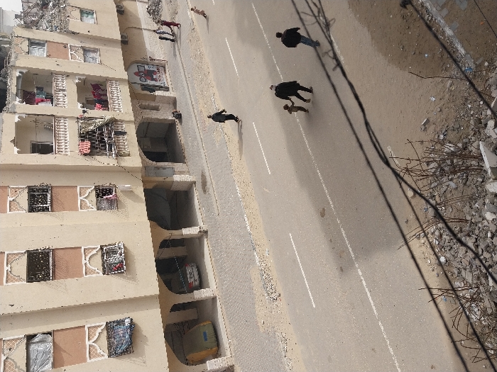 This image presents an elevated view of a street in Khan Yunis, Palestinian Territory. The scene is dominated by a multi-story, light-beige building featuring decorative window frames and multiple balconies, some displaying hung laundry. The upper right section of the building shows significant structural damage, including exposed rebar and rubble from a partial collapse. The ground floor incorporates arched open bays; a white sedan is partially visible within one, and a yellow-wrapped object in another on the far left. Red and black graffiti marks a wall segment on the ground level near the damaged part of the building.
The asphalt street, featuring faded white lane markings, is bordered by unpaved shoulders with scattered debris. The foreground consists of substantial concrete rubble and exposed rebar.
Multiple individuals are present: a lone person in dark clothing walks leftward on the asphalt. To their right, two adults and a small child holding an adult's hand walk away from the viewer. On the right side of the street, near the building, a group of four individuals—comprising two children, an adult in a red garment, and another person—is visible. A fifth child in a light top moves quickly on the unpaved shoulder further right.