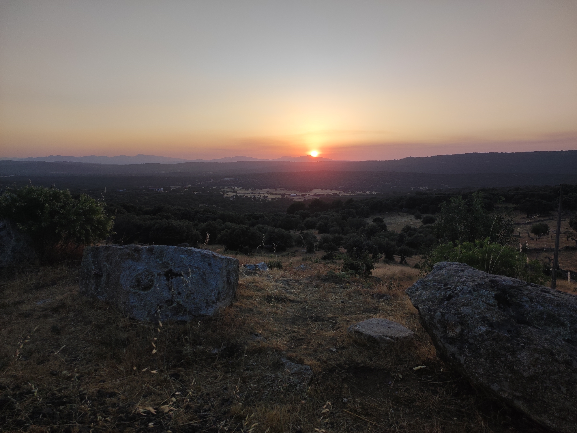 This is a wide-angle, landscape photograph of a sunset over a rural area, likely in Ledrada, Spain. The foreground is dominated by two large, rough-textured boulders, one on the left and one on the right, with dry, straw-like vegetation scattered around them. In the midground, a dark, undulating expanse of trees, possibly olive groves, stretches towards the horizon. Beyond the trees, a valley is visible with some scattered buildings and fields, bathed in the soft, warm light of the setting sun. The distant background is a range of low, hazy mountains silhouetted against the sky. The sky itself transitions from a pale yellow and orange at the horizon where the sun is setting, to a soft pink, and then to a light greyish-blue at the top of the frame. The sun, a bright orange disc, is partially obscured by the horizon line. There are no people or animals visible in the image. The overall impression is one of serene, natural beauty at dusk.