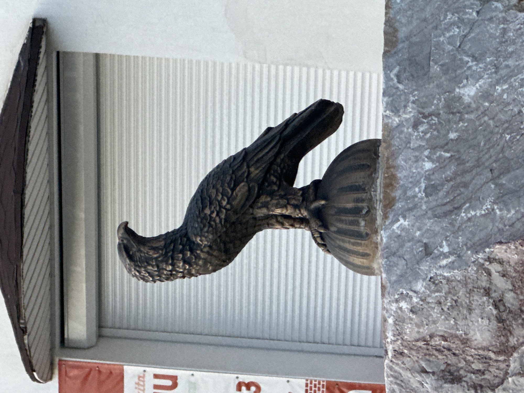 A metal sculpture of an eagle in profile, possibly bronze, is mounted to a building facade. The building has a vertically ribbed metal cladding section directly behind the eagle. To the right of the eagle, there is a rough, potentially stone or concrete structural element with a marbled texture. Below the eagle and cladding, a small section of a red and white banner with text is visible. The setting appears to be urban, as indicated by the commercial signage. Location details are not discernible from the image.