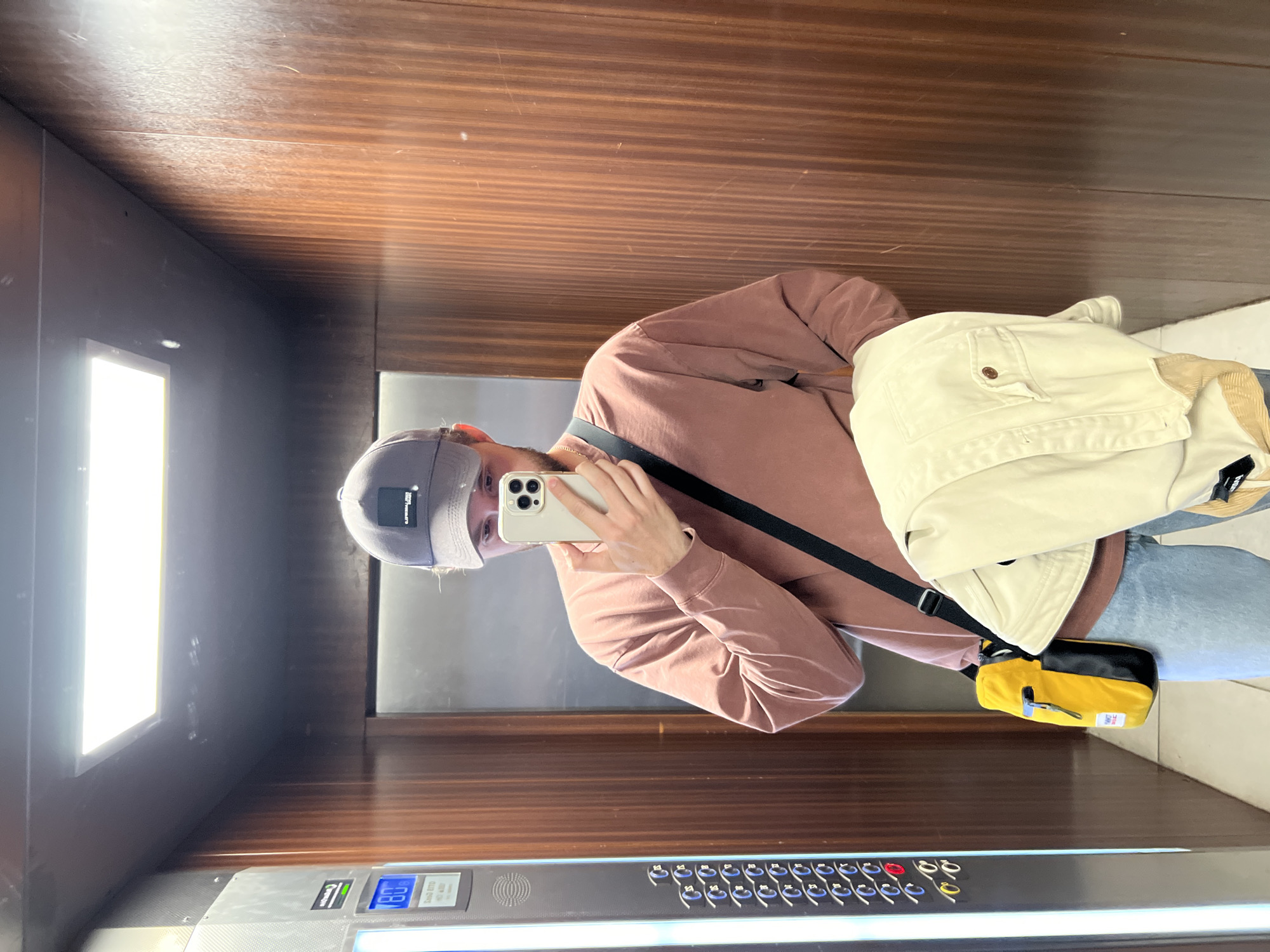 A young man wearing a pink long-sleeve shirt, light blue jeans, and a grey baseball cap is taking a mirror selfie in an elevator. He is holding a smartphone in his right hand. A yellow and black crossbody bag is slung over his left shoulder. A cream-colored jacket is draped over his left arm. The elevator has wood-paneled walls, a metal control panel with buttons and a digital display showing the number 80, and a fluorescent light fixture on the ceiling. The image appears to be taken in an urban environment in Krasnoyarsk, Russia, based on the elevator's design which is typical of many residential buildings in Russian cities.