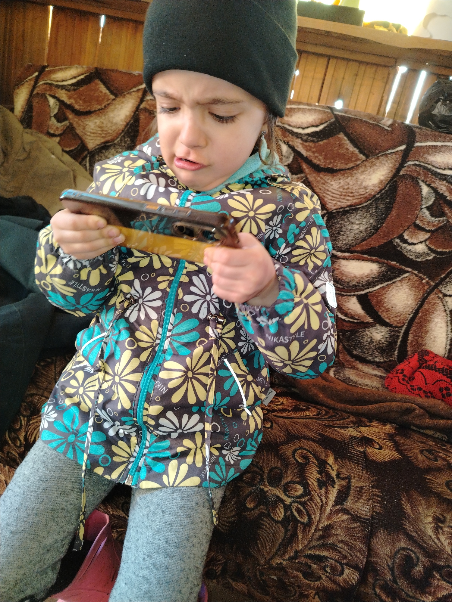 A young child with a black beanie sits on a brown patterned couch and holds a smartphone in their hands. The child wears a floral-patterned jacket and gray leggings. The background includes a wooden wall and other items on the couch. The location is not discernible from the given information.