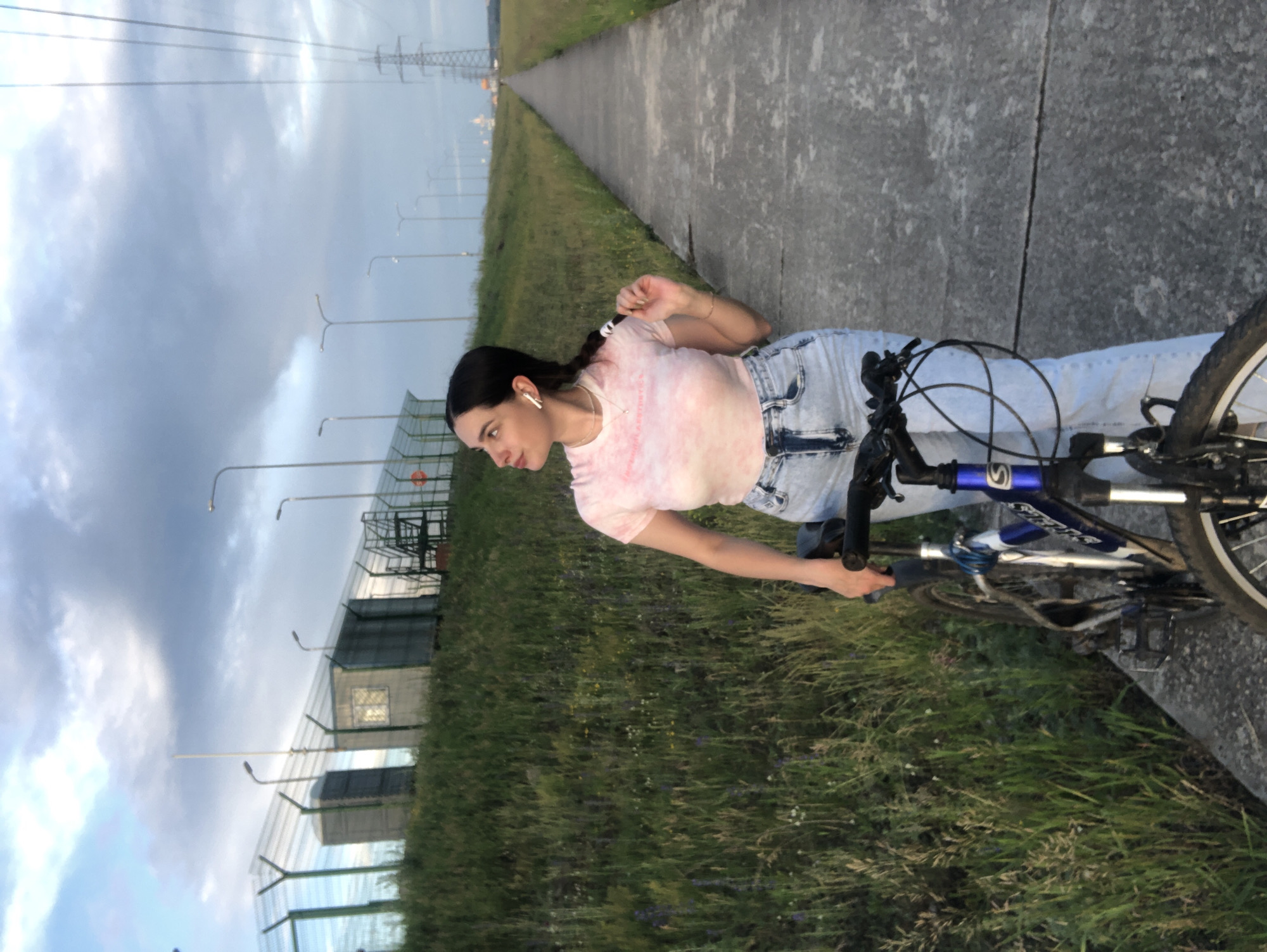 A young woman stands on a paved pathway next to a bicycle. She is wearing a light pink tie-dye t-shirt, light wash high-waisted jeans, and white earbuds. Her hair is dark and braided, and she is looking up and to her left, touching her braid. The bicycle is dark blue and silver. The pathway runs alongside a grassy embankment on the left and continues into the distance on the right. In the background, there is a fence, several small buildings, streetlights, and a large power line tower. The sky is cloudy with patches of blue. The scene appears to be in an open, semi-industrial area on the outskirts of Noviy, Russia.
