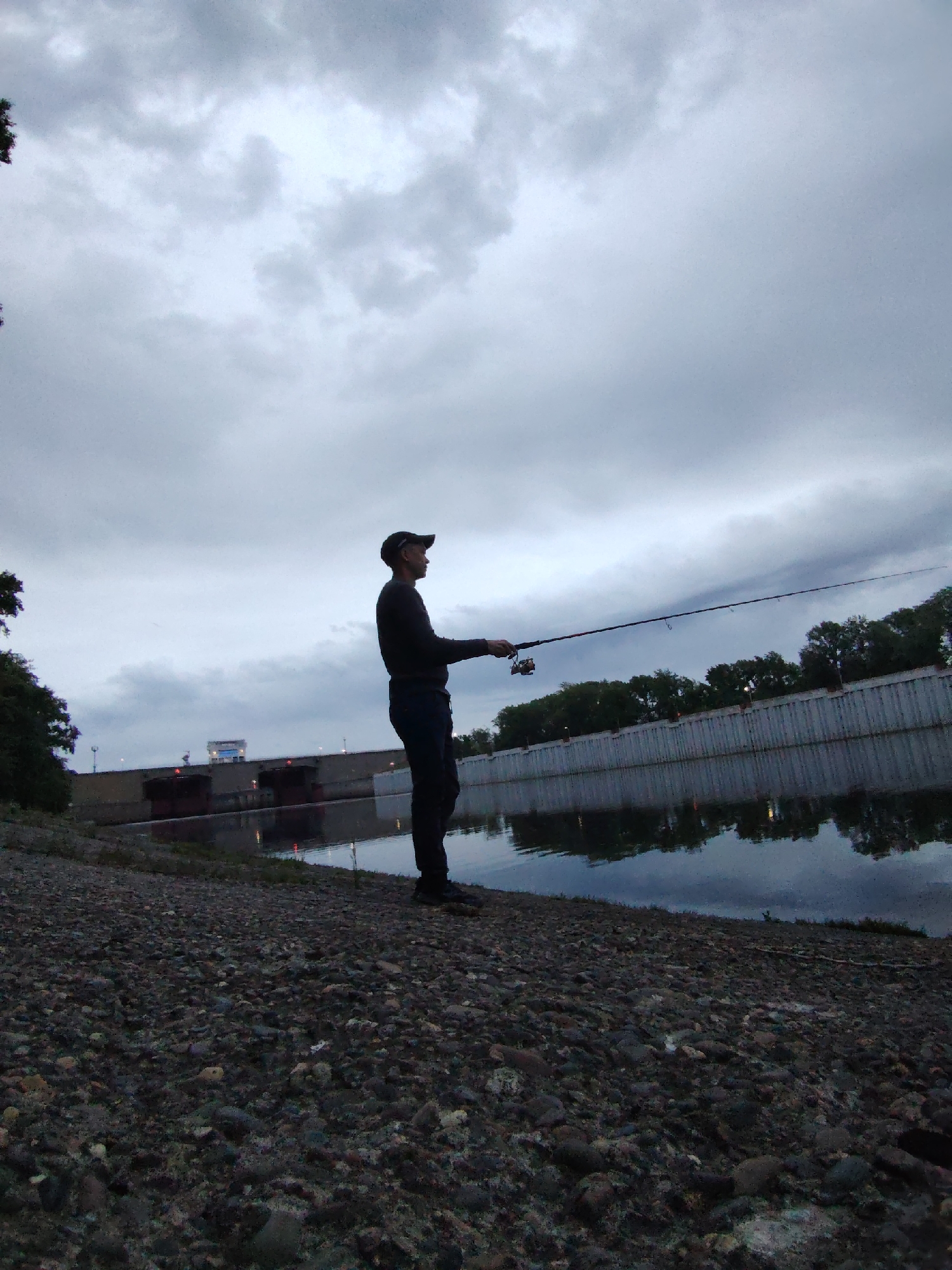 A man stands on a rocky embankment, fishing in a body of water on an overcast day. The man, silhouetted against the light sky, wears a cap, long-sleeved shirt, and dark pants. He holds a fishing rod extended over the water. Behind him, a concrete embankment lines the water, with trees visible along the top of the embankment. In the distance, a structure resembling a dam or bridge is visible. The scene appears to be in Chaikovsky, Russia, based on visual features of the landscape and possible water structures. The ground is covered with dark-colored stones and gravel, suggesting a riverbank or shoreline.