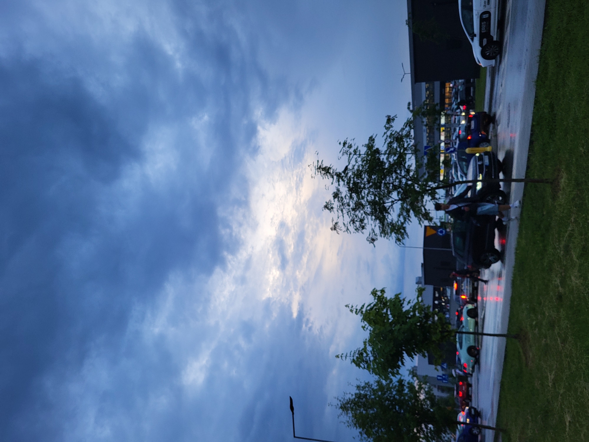 The image depicts an urban or suburban street scene taken at what appears to be dusk or dawn, with a prominent cloudy sky. The ground is wet, suggesting recent rain.
The sky occupies a large portion of the frame, filled with dense, blue-grey clouds. A brighter, lighter patch of sky is visible towards the center-right, indicating a light source (likely the sun) behind or breaking through the clouds.
At ground level, a wide paved area, possibly a road or parking lot, is visible, lined with a strip of green grass on the right. Several dark green trees are scattered along the right and lower-left edges, some partially obscuring the view. Multiple cars are present; some appear parked while others might be in traffic, with their headlights and taillights glowing, reflecting on the wet surface. A white car, possibly a taxi or ride-share vehicle, is prominently seen in the upper right, bearing text that includes "Uber" and "SP2S." Other vehicles are darker, including blue, green, and black cars.
In the background, dark, modern-style buildings with lit windows are visible. A few human figures can be seen near the cars; one person in a light top and dark shorts is noticeable towards the center-bottom, appearing to walk or stand. A dark street light pole stands in the lower-left corner. The overall scene captures a moment of ordinary urban activity under a dramatic, cloudy sky.
