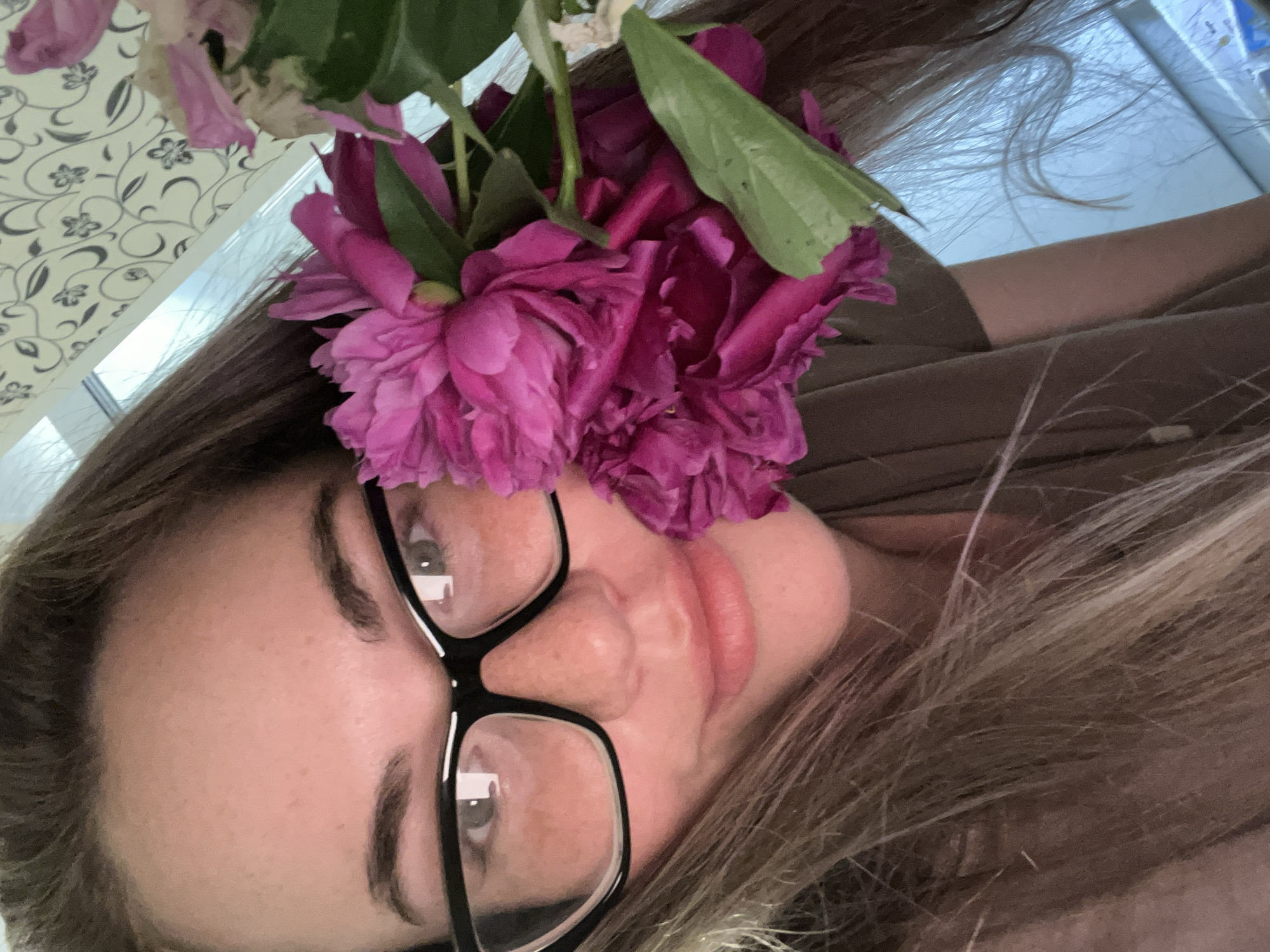 A close-up, high-angle, medium shot shows a Caucasian woman with long brown hair, wearing black-rimmed glasses, and a brown t-shirt. She is partially obscured by a bouquet of bright pink peonies and green leaves. The background features a light-colored wall with a subtle floral wallpaper pattern and a window with a view of a bright, overcast sky. The overall lighting is soft and natural, suggesting an indoor setting. No specific location in Chaikovsky, Russia, can be identified from the image.
