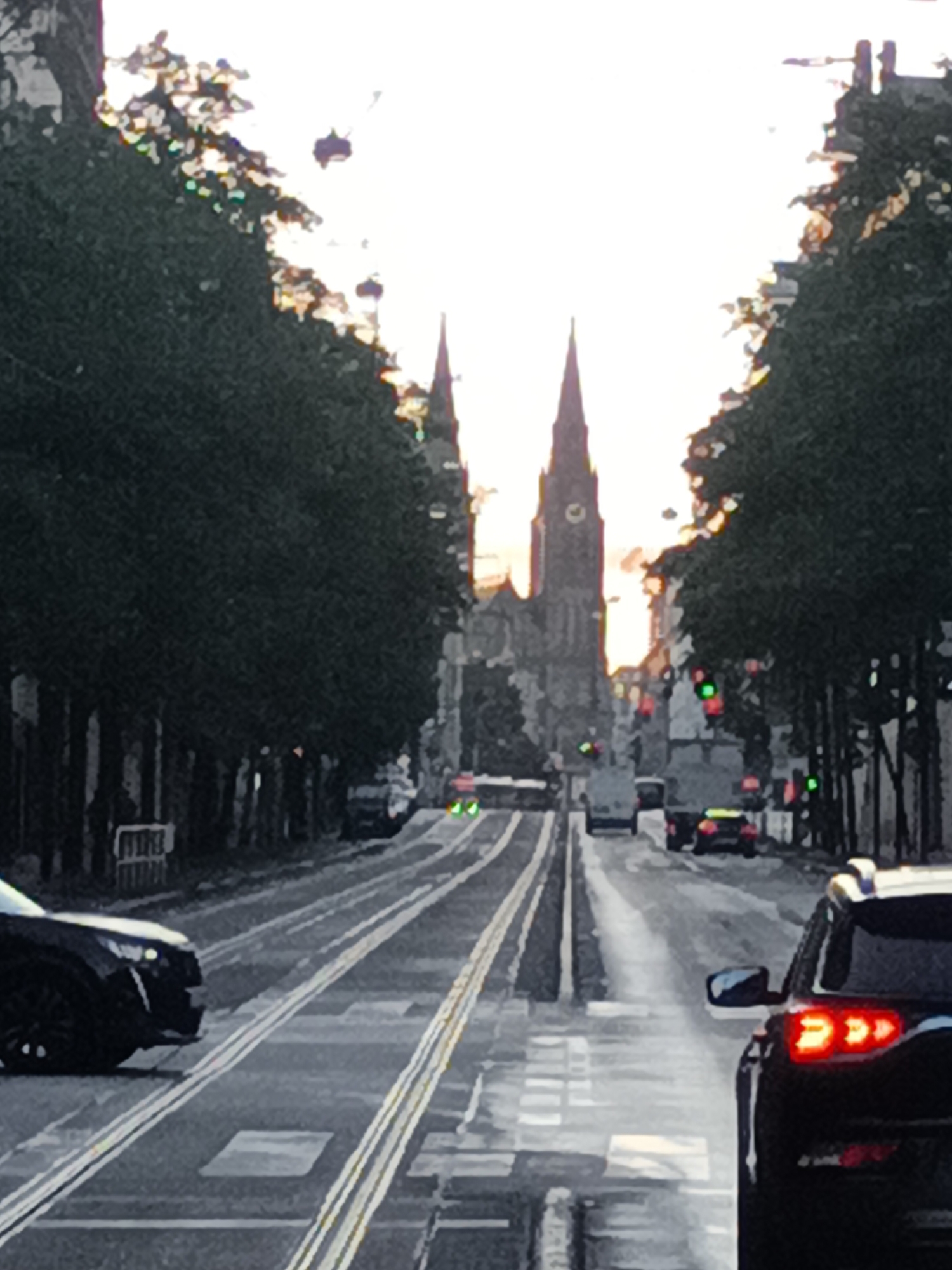 The image shows a view down a city street with tram tracks in the foreground. Two large church spires, likely part of the St. Vitus Cathedral in Prague, Czechia, are visible in the distance. Trees line both sides of the street, and several cars are driving on the road. The sky is bright and hazy, suggesting either sunrise or sunset.