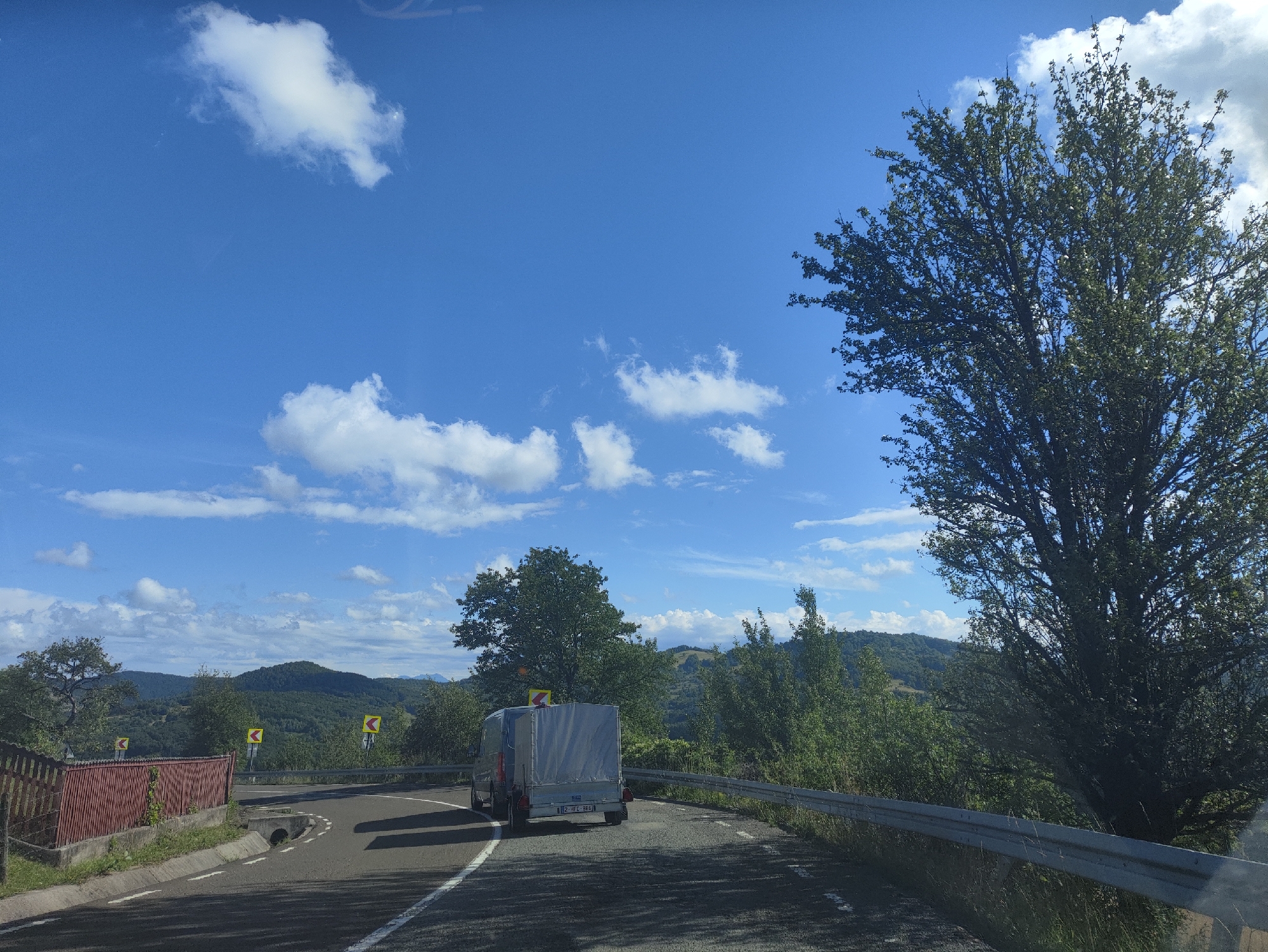 This media file depicts a scenic drive through a mountainous, rural landscape in Petrova, Romania, on a bright, sunny day. The scene is captured from the perspective of a vehicle traveling on a winding asphalt road.
The foreground features the road itself, with white lane markings and a concrete drainage ditch. To the left, a red wooden fence lines the side of the road, adjacent to a grassy embankment. A large, leafy tree dominates the right side of the frame, its branches reaching towards the vibrant blue sky dotted with fluffy white clouds. In the distance, rolling hills covered in dense green forests create a picturesque backdrop.
A small, silver van with a white tarp-covered trailer is visible on the road, moving away from the viewer and around a curve. Several yellow road signs with black arrows indicating sharp turns are strategically placed along the roadside, suggesting the challenging nature of the route.
The overall impression is one of a pleasant journey through a beautiful natural environment. The clear sky and lush greenery suggest it is daytime, likely late morning or early afternoon. The weather appears to be warm and fair, ideal for driving and enjoying the scenery. The presence of the road, fence, and road signs indicates a developed, albeit possibly remote, area. The license plate on the trailer is partially visible, showing "12 JFC 868". There are no people clearly visible in the image.