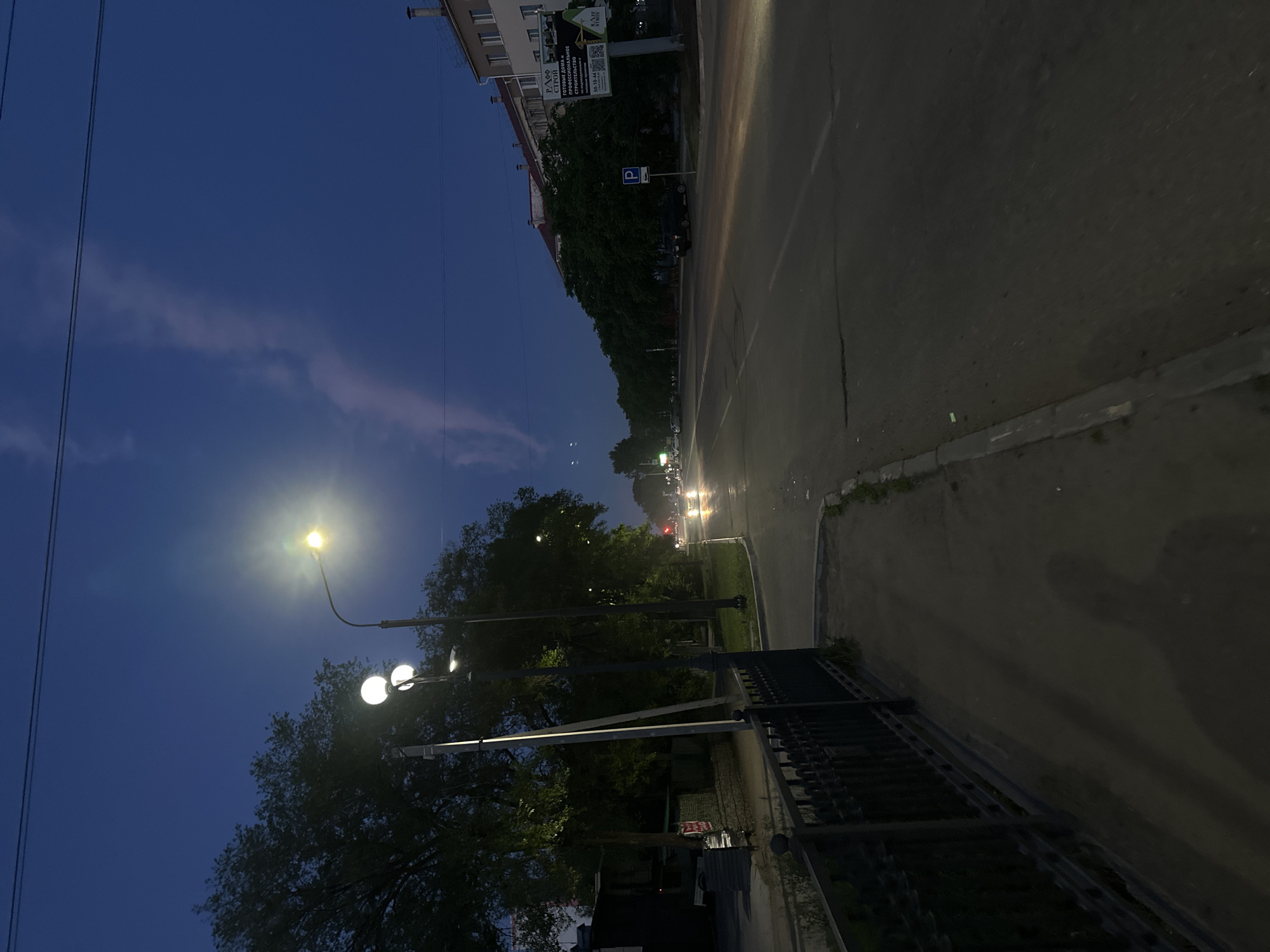 The image depicts a street scene at dusk in Blagoveshchensk, Russia. A paved road with faint lane markings stretches into the distance. On the left, a sidewalk runs alongside a black metal fence and a row of trees. Streetlights are illuminated, casting a glow on the surroundings. Several cars are visible on the road, with their headlights on, suggesting approaching vehicles. A building with multiple windows stands on the right side of the street, and a billboard with Cyrillic text is mounted on its side. The sky is a deep blue with a faint, wispy cloud formation. The overall impression is that of a quiet urban evening.