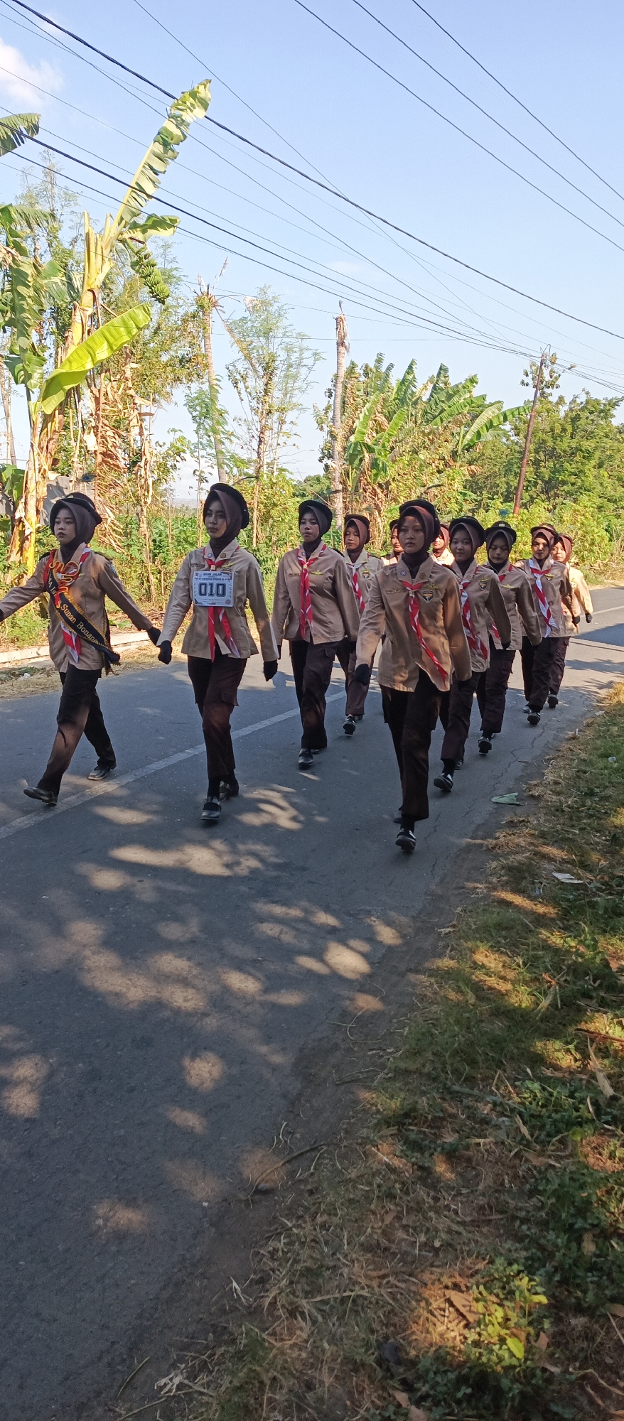 In Ketowan, Indonesia, a group of approximately ten young women, dressed in scout uniforms consisting of brown shirts, brown pants, and brown headscarves, are marching in single file on the right side of an asphalt road. They are wearing red and white neckerchiefs. The lead scout on the left has a sash draped across her chest with text that is partially obscured. The second scout from the left is wearing a bib with the number "010" and some Indonesian text, indicating a competition or event. The background shows a road with a white line marking the edge of the lane, trees lining the left side of the road, and a clear blue sky above. Sunlight casts shadows of the trees onto the road, suggesting a sunny day. The environment appears rural or suburban.