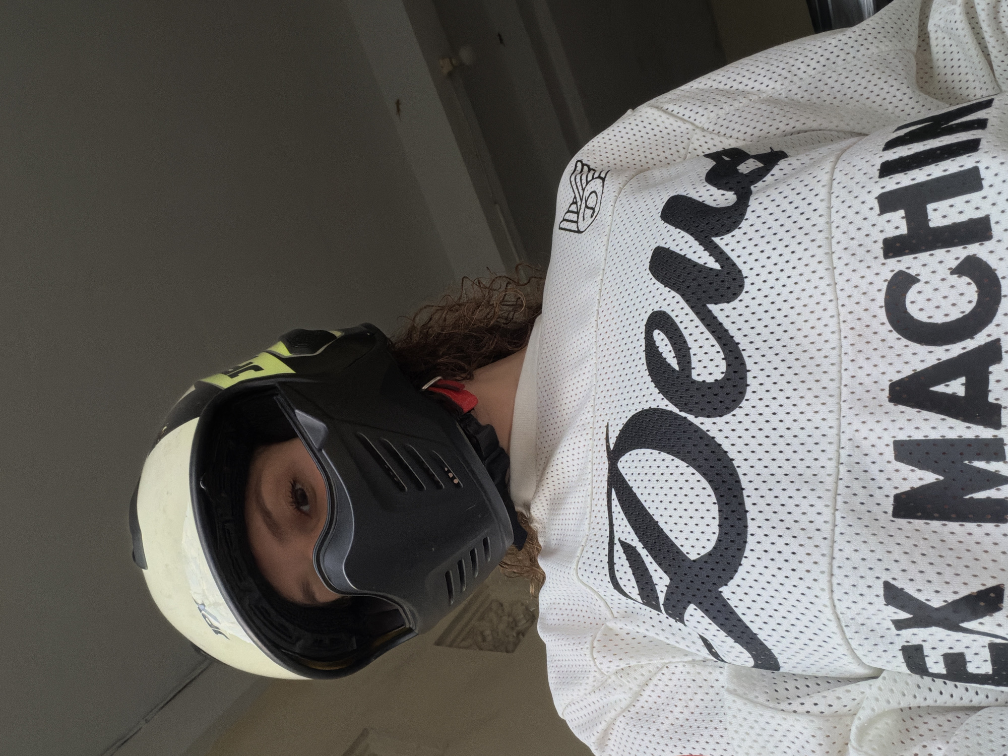 A person wearing a white and yellow motorcycle helmet and a white mesh jersey with black lettering is seen from the chest up, looking directly at the camera. The jersey has the words "Deus Ex Machina" printed on it in large black letters. The background is dimly lit and appears to be indoors, possibly a garage or covered area. The image does not provide any specific information about the location in North Kuta, Indonesia.