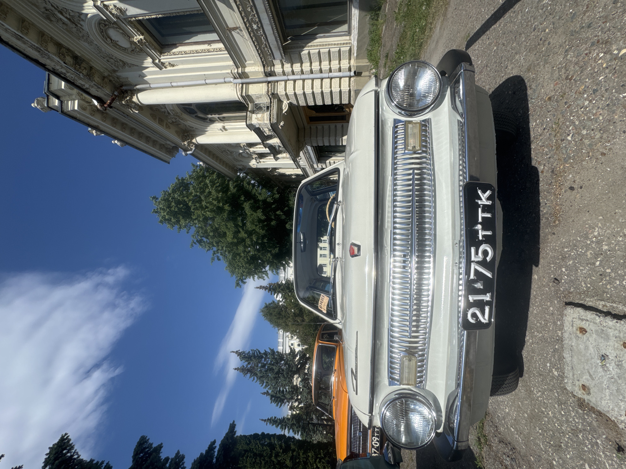 The image shows the front of a white vintage car with the license plate "2175 TTK" parked on an asphalt street in front of an ornate, light-colored building. A second orange car is partially visible behind the white one. The sky is blue with scattered clouds. Trees and a building are also visible in the background.