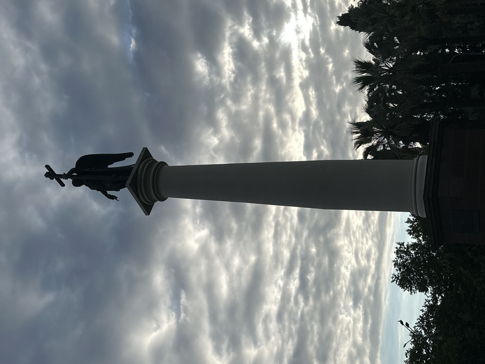 A silhouette of an angelic statue holding a cross atop a tall, classical column stands against a cloudy sky. The statue depicts an angel with outstretched wings, its gaze seemingly directed forward. The column is a light color and features a decorative capital. Below the column is a dark, rectangular base, partially obscured by trees. In the background, dark silhouettes of palm trees and other foliage are visible. The sky is filled with dramatic, grey and white clouds, suggesting overcast weather. This monument is located in the city of Sochi, Russia, specifically on a hill overlooking the Black Sea, known as Kolyada Hill. It is part of the Alexander Nevsky Cathedral complex.