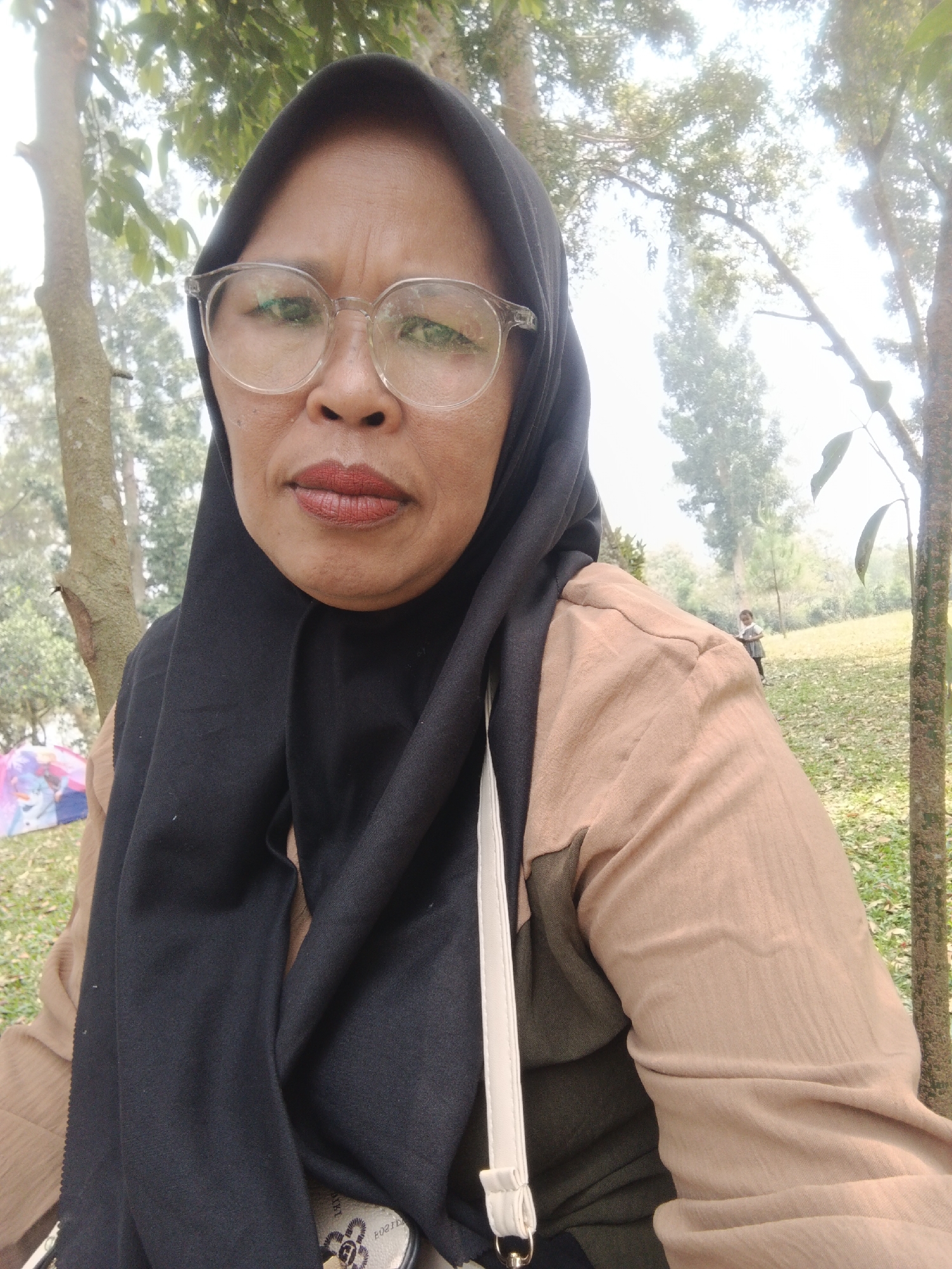A selfie is taken by a woman wearing a black hijab and glasses, with reddish lipstick, in an outdoor setting. She is wearing a tan shirt and a white strap is visible across her chest. In the background, a child is walking up a grassy hill, and a pink and blue tent is partially visible to the left. The environment appears to be a park or wooded area with trees and fallen leaves. The location is identified as Perbawati, Indonesia.