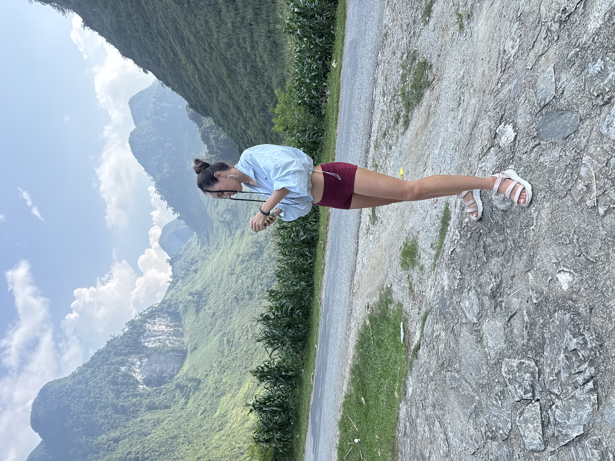 A woman stands on a paved area in Đường Thượng Commune, Vietnam, with mountains and vegetation in the background. She has her hair in a bun, wears a light blue, button-down shirt and maroon shorts, and pink sandals. She looks down, holding something in her hands. The surrounding environment includes a paved road, a field of crops, and a sky with light clouds.