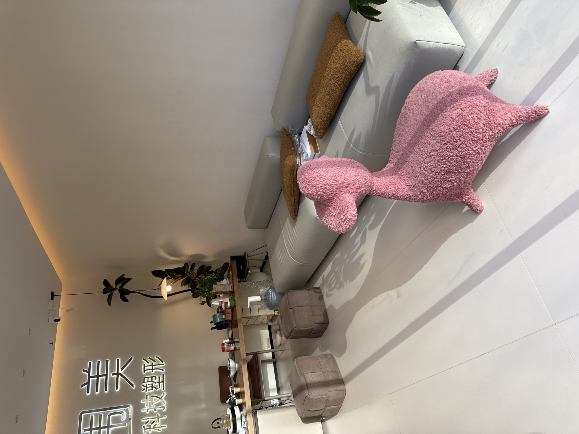 This image depicts a modern interior space, likely an office or waiting area, located in the city of Yiwu, China. The room is brightly lit, with natural light casting distinct shadows across the white tiled floor from an unseen window.
Dominating the foreground is a plush, pink, animal-shaped ottoman, possibly a decorative chair, positioned on the floor. Behind it, a long, light gray modular sofa is arranged along the wall. On the sofa, two textured, brown rectangular cushions are placed, along with a white box and some books.
To the left of the sofa, two square, brown upholstered ottomans are situated on the floor. Further to the left, a wooden desk or counter is visible, cluttered with various items including a kettle, bowls, bottles, and what appears to be a water dispenser. Two chairs are placed at the desk.
The wall in the background features illuminated neon signage in Chinese characters, adding a contemporary aesthetic. Above the signage, a plant is suspended, and a pendant light fixture hangs from the ceiling. A small security camera is also mounted on the ceiling. The overall environment suggests a clean, minimalist, and well-lit commercial or public space.