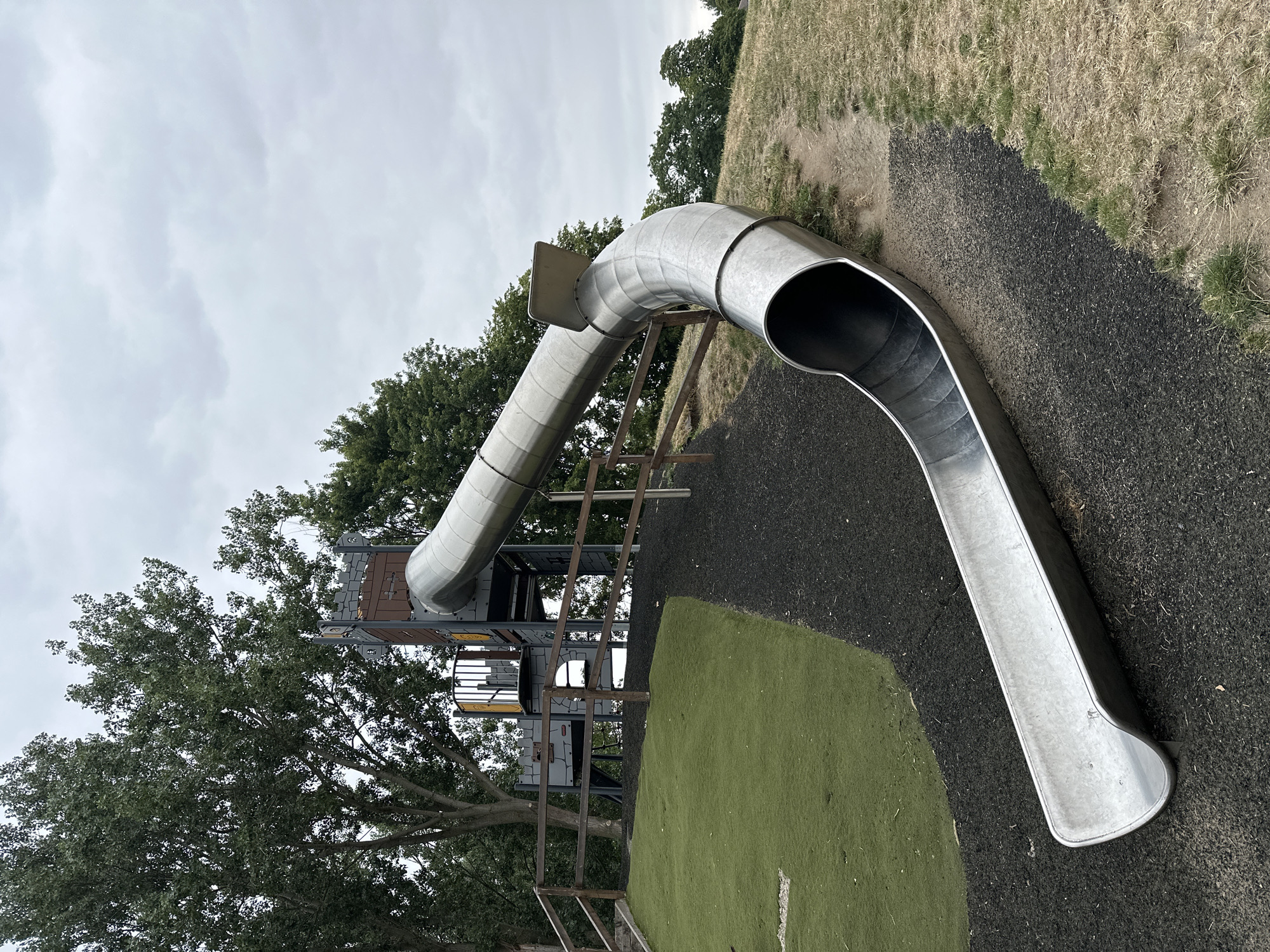 This image shows a playground slide with a castle-themed structure at the top, located in the city of Wakefield, United Kingdom. The slide is made of metal and curves downwards onto a rubber safety surface. To the left of the slide, there is a grassy hill with a wooden fence. Trees and a cloudy sky are visible in the background. The scene is an outdoor public space.