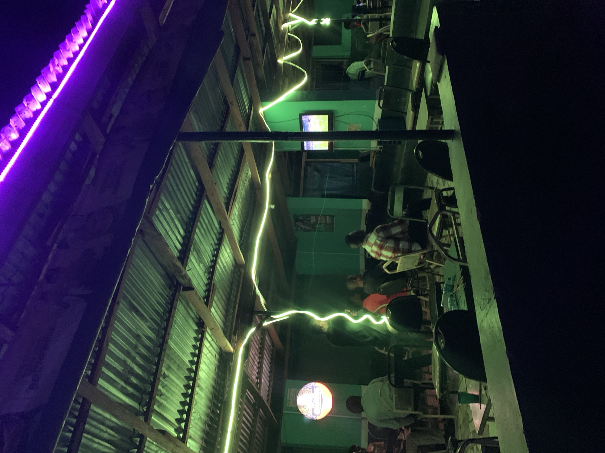 The image depicts an outdoor seating area, possibly a restaurant or bar, in Jos, Nigeria, at night. The space is illuminated by a combination of lighting, including a bright purple light strip along the edge of the roof, a string of yellow lights running the length of the ceiling, and a round decorative light.
Several people are present, seated at tables and chairs. One person is wearing a red and white plaid shirt. A television screen is visible on a wall, displaying an image. The roof is made of corrugated metal sheets. The walls are painted green. The furniture appears to be simple and functional. The overall atmosphere seems relaxed and social.