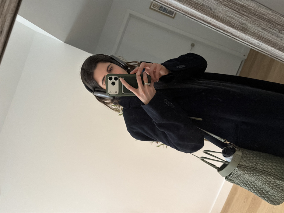 The image displays a female subject taking a mirror selfie in an interior space within New York City, United States of America. The subject, visible from the mid-torso upwards, has dark hair and wears grey over-ear headphones and a dark, long coat. She holds a smartphone with a green protective case, featuring a visible triple-lens camera array and a dark accessory, possibly a card holder, attached to its back. Her left hand grips the phone, partially obscuring her face, while her right hand, adorned with blue nail polish and a ring on the fourth finger, supports the phone from below. A light olive green woven-texture tote bag with two top handles and a shoulder strap hangs from her left shoulder, containing a dark rolled item. The background, reflected in the mirror, consists of plain white walls, light wooden flooring, a white door with a silver handle, and a small framed object on the far right wall. The mirror itself has a light-colored, textured wooden frame. No specific street or architectural location within New York City is discernible from the visual information provided.