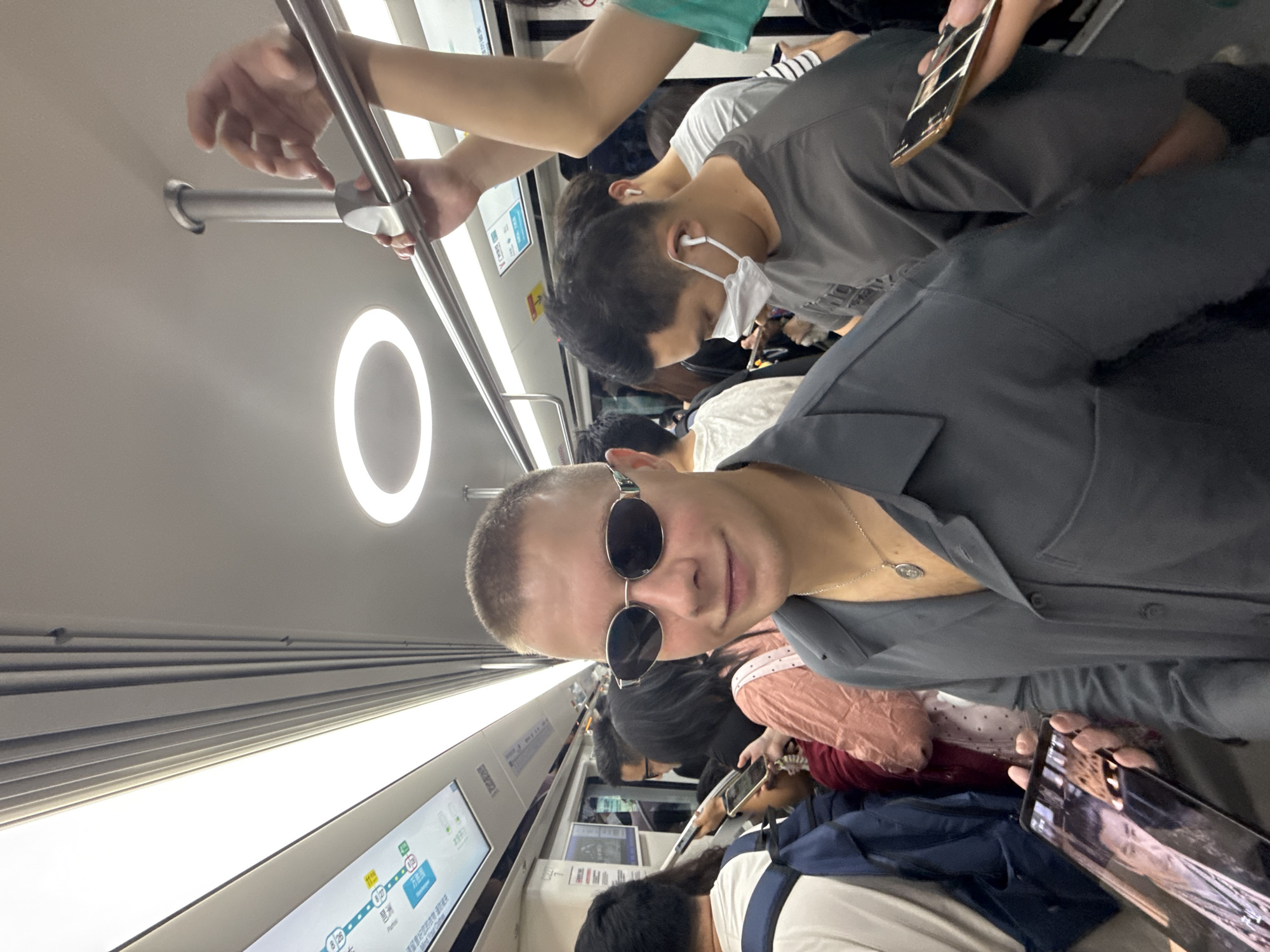 A young Caucasian male with a buzz cut and sunglasses is posing for a selfie on a crowded subway train. He is wearing a grey collared shirt and a silver chain. In the foreground, he is holding a smartphone that displays an image of another young man. Behind him, several other passengers are visible, some looking at their phones, others wearing masks and earbuds. The subway car appears to be in motion, with overhead handles and lights indicating an urban transit environment. The image was taken in the Haizhu District of Guangzhou, China.