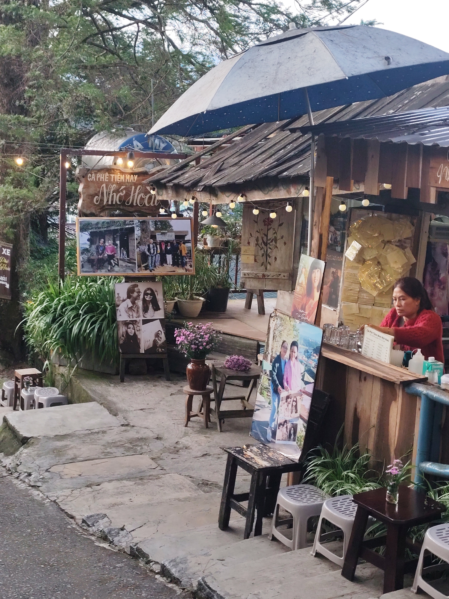 The image depicts an outdoor cafe in Da Lat, Vietnam, identified by a sign that reads "CÀ PHÊ TIỆM HAY Nhớ Hoa". The cafe is situated on a stepped, sloped pathway, suggesting a hillside location common in Da Lat. A woman, wearing a red sweater, is behind a wooden counter, appearing to serve or prepare something. She is facing towards the right side of the frame.
In front of the cafe, several photographs are displayed, including larger framed prints and smaller mounted pictures. One large photo collage shows groups of people in outdoor settings. Another large print displays two women, and a smaller display features a man and a woman, and another of a woman.
The cafe structure has a rustic appearance with a corrugated metal roof and wooden walls. An open umbrella, partially covering the roof, is positioned above the seating area. String lights are draped across the front of the cafe, adding to the ambiance.
Various seating options are present, including small wooden stools and plastic stools arranged on the steps. A vase with pink flowers sits on a small wooden table. Lush green plants are strategically placed around the cafe, contributing to the natural environment. The pathway is made of concrete slabs, showing signs of wear and cracking. In the background, dense foliage and trees are visible, indicating a verdant setting typical of Da Lat.