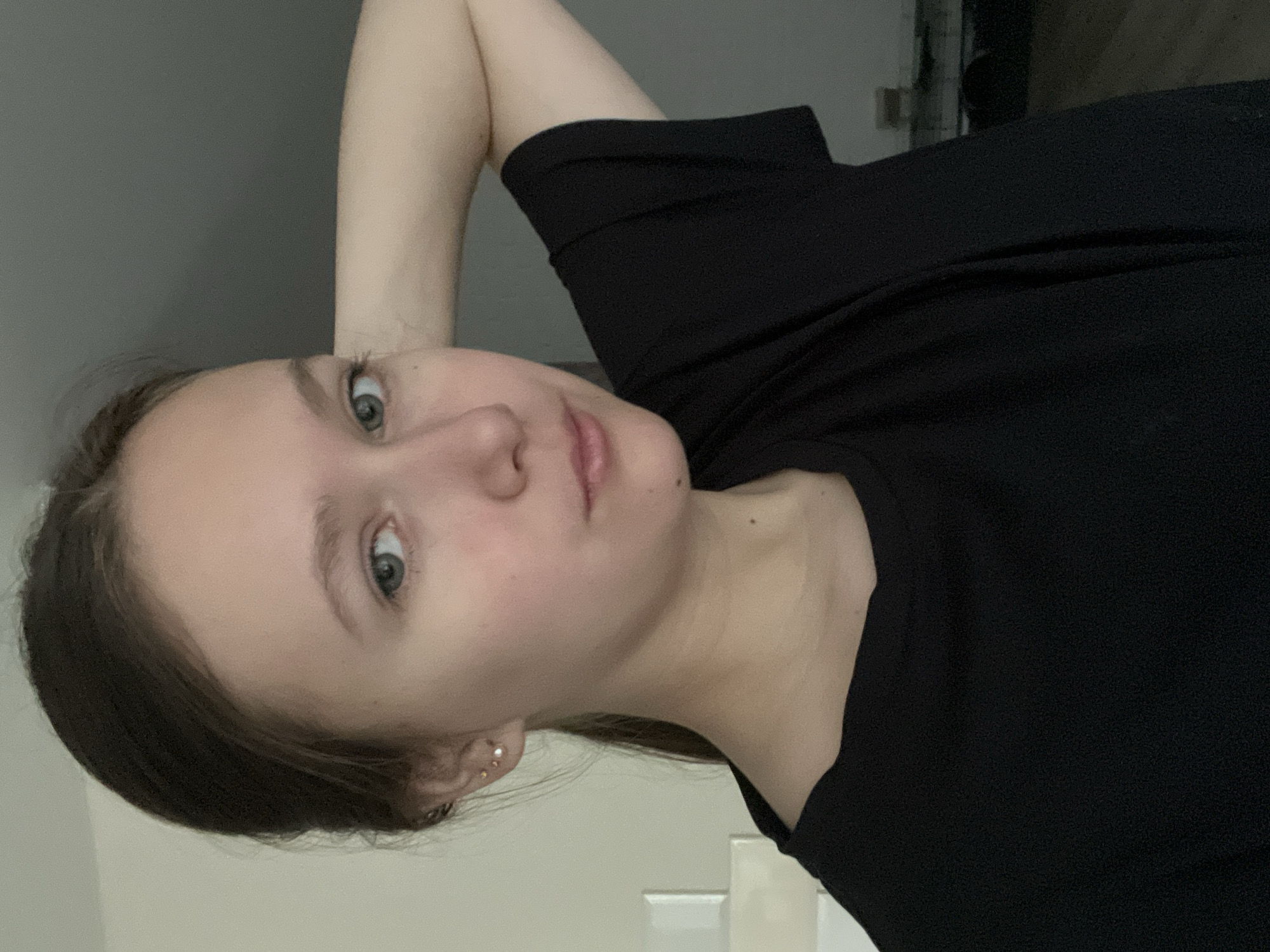 The image depicts a young adult female in an indoor setting. She has fair skin, dark brown hair styled in a low ponytail, and prominent greenish-blue eyes that gaze directly forward. Her expression is neutral, with her lips slightly parted. She is wearing a simple black short-sleeved t-shirt. On her visible left ear, three small stud earrings are worn: a gold-colored sphere, a white pearl-like sphere, and a smaller silver-toned stud higher up. Her left arm is raised, with her hand positioned behind her head or neck, giving a relaxed posture. The background on the left side of the frame is a light, off-white wall with two rectangular white ceramic tiles or raised molding visible behind her shoulder. The background on the right is a darker gray wall or surface, appearing more shadowed. The lighting is soft and even, illuminating her face. The image itself contains no discernible landmarks or contextual elements that specify its location within the city of Chaikovsky, Russia.
