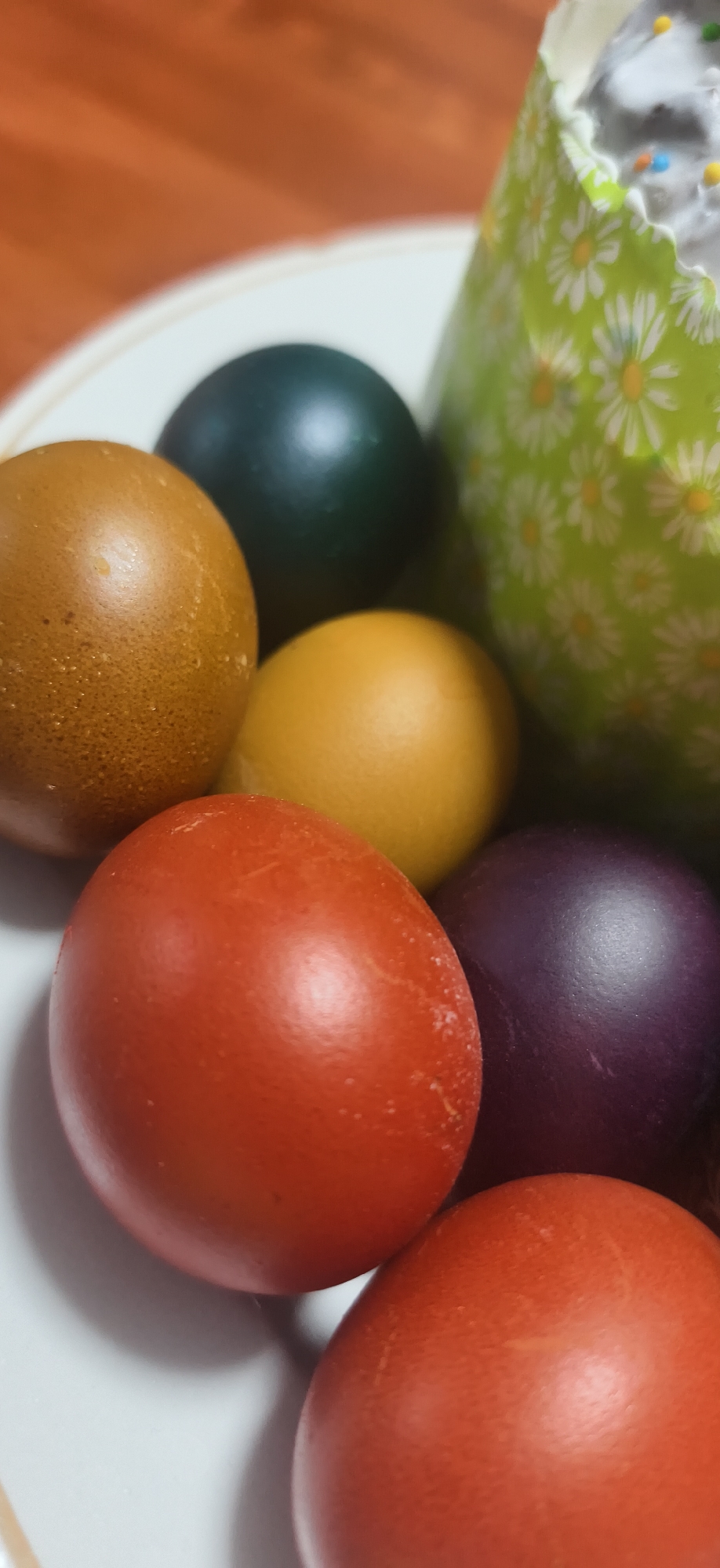 The image shows a close-up of several colored eggs in a bowl, next to what appears to be an Easter cake decorated with white flowers and sprinkles. The eggs are various colors, including brown, green, yellow, red, and purple. The scene is indoors. There are no people visible. There is no evidence to confirm the image's location is Фоки, Russia.