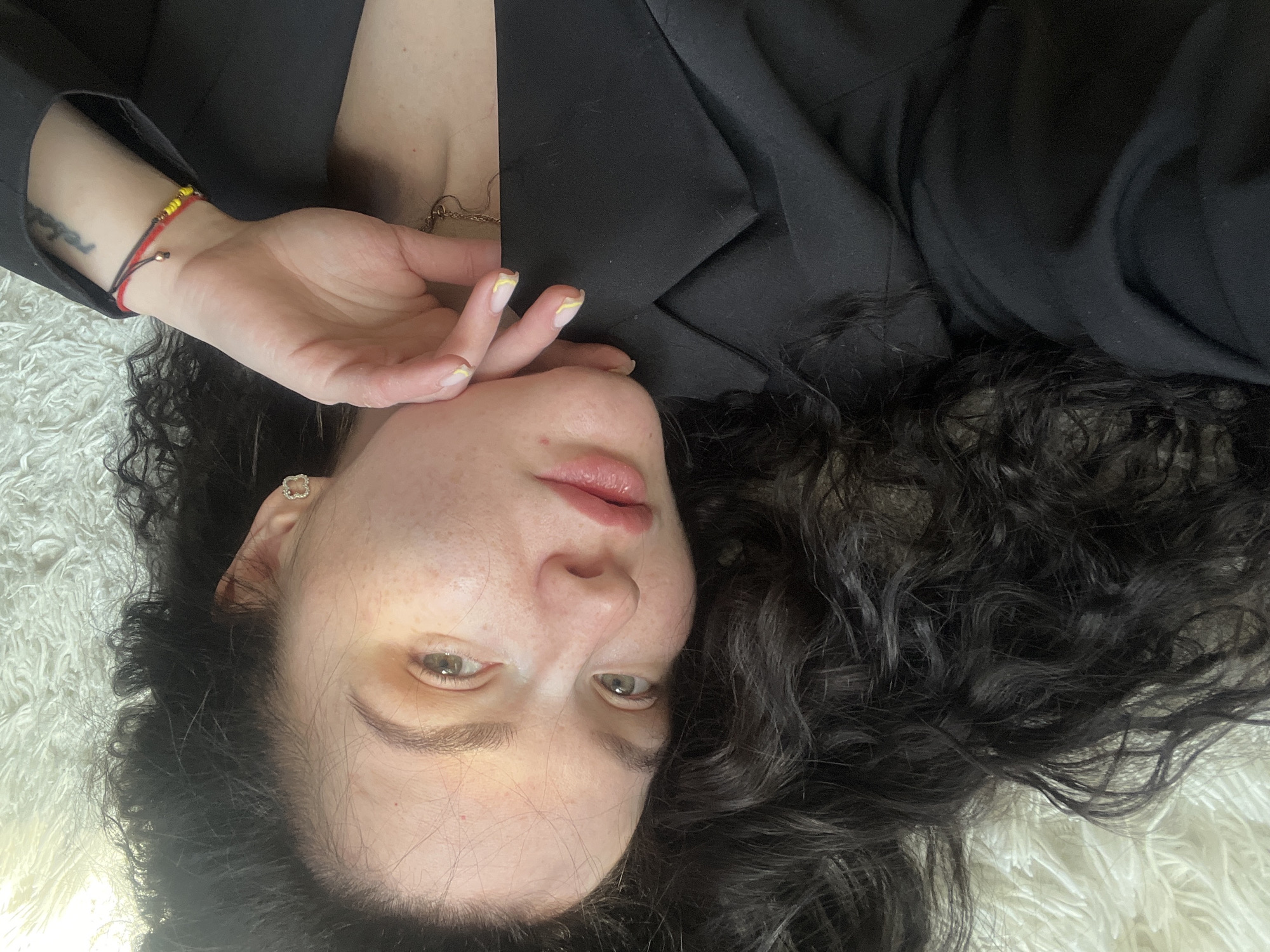 A young woman with fair skin, freckles, and dark curly hair lies on a white fluffy surface, possibly a rug. She is wearing a black blazer. Her left hand is near her face. Her nails are painted with a pale yellow accent. She wears a beaded bracelet on her left wrist. The setting appears to be indoors. The location of the scene is not determinable from the image.