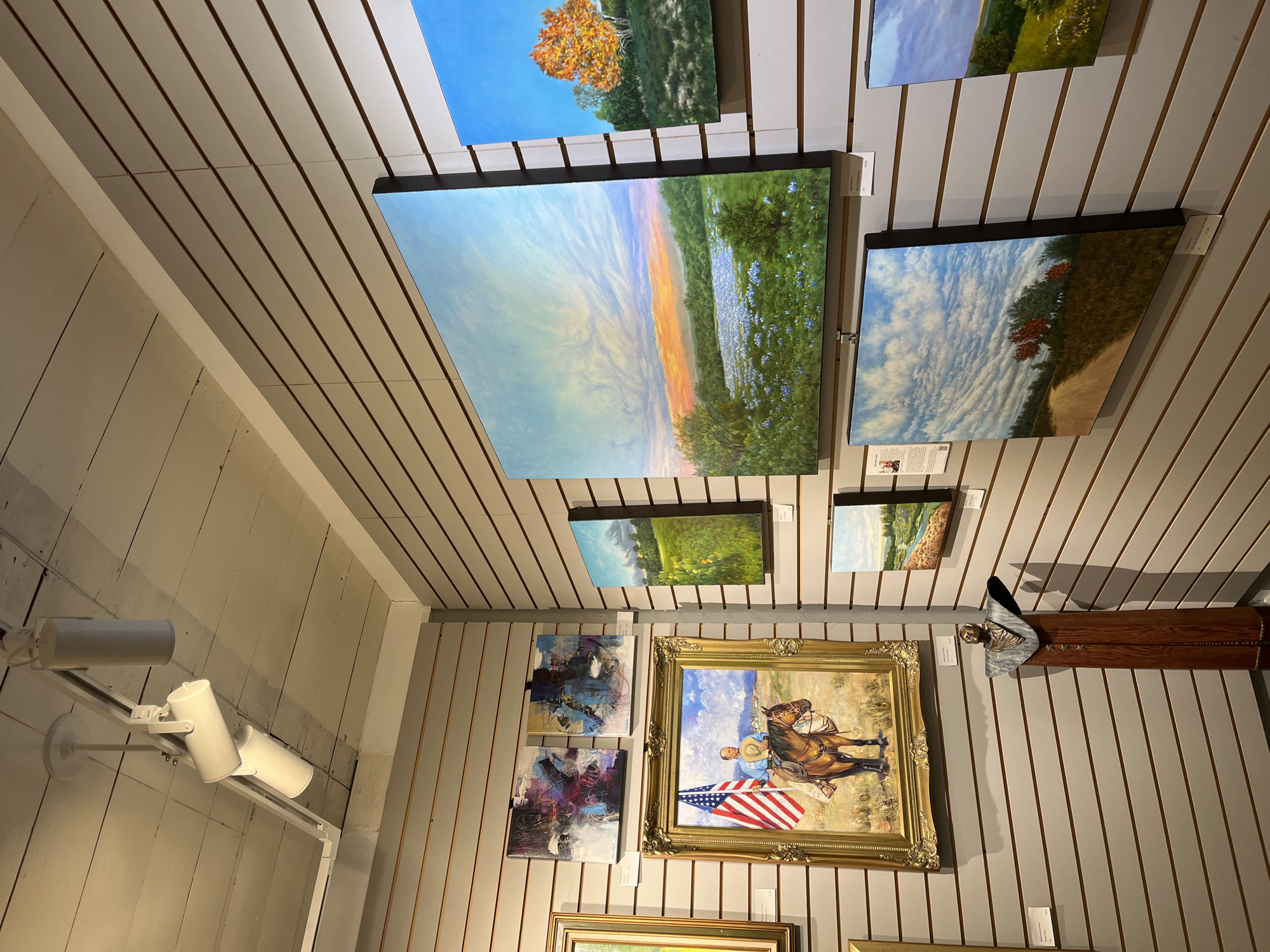 The image shows an interior space, likely an art gallery or shop in Wimberley, Texas. The walls are covered in a horizontal striped pattern with several framed paintings of landscapes, abstracts, and portraits are hung. At the left of the image are white adjustable spotlights mounted on the wall. The paintings vary in size and style, and include landscapes with trees, flowers, and skies, an abstract piece, and a portrait of a man on horseback holding an American flag, framed in gold. The lighting is mixed, with both natural light and artificial light from the spotlights illuminating the scene. The gallery appears to be well-maintained and professionally presented.