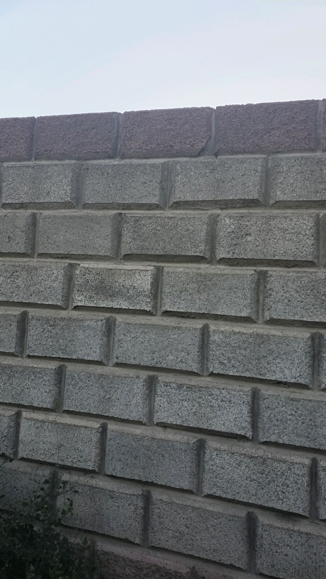 The image shows a close-up, low-angle view of a gray brick wall with a darker colored capstone row at the top. The wall is constructed of rectangular concrete blocks with a textured surface, laid in a stretcher bond pattern. The sky is visible at the top of the image, appearing overcast and light in color. Some greenery is visible in the lower left corner of the frame. The scene is devoid of any people or animals. The location is identified as Chuy, Kyrgyzstan.