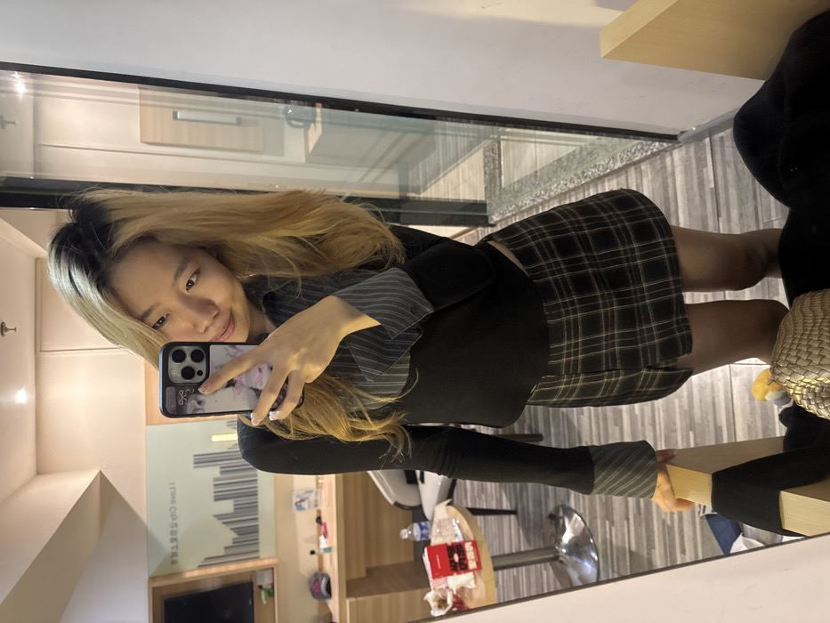 An East Asian female is depicted standing indoors, taking a mirror selfie in what appears to be a hotel room or residential unit located within Yuzhong District, China. She has long, blonde hair with visible dark roots and is wearing a black long-sleeved top with striped grey and white collar and cuffs, paired with a black and brown plaid mini-skirt. She holds an iPhone, equipped with a patterned case, in her right hand, positioned to capture her reflection. Her left hand rests on a light-colored wooden furniture edge in the foreground.
The room's reflection in the mirror reveals a light-colored wall adorned with a framed graphic featuring a cityscape silhouette and text, partially legible as "I love Chongqing." Below this, a wooden desk holds a red baseball cap, small bottles, and a clear water bottle. To the left, a small round table with a metallic base supports a red cigarette packet and another clear water bottle. A white chair with a metallic base is partially visible behind the table. The floor appears to be light-colored, possibly tiled or laminated. To the right of the subject, a tall glass panel or mirror reflects further into the room.