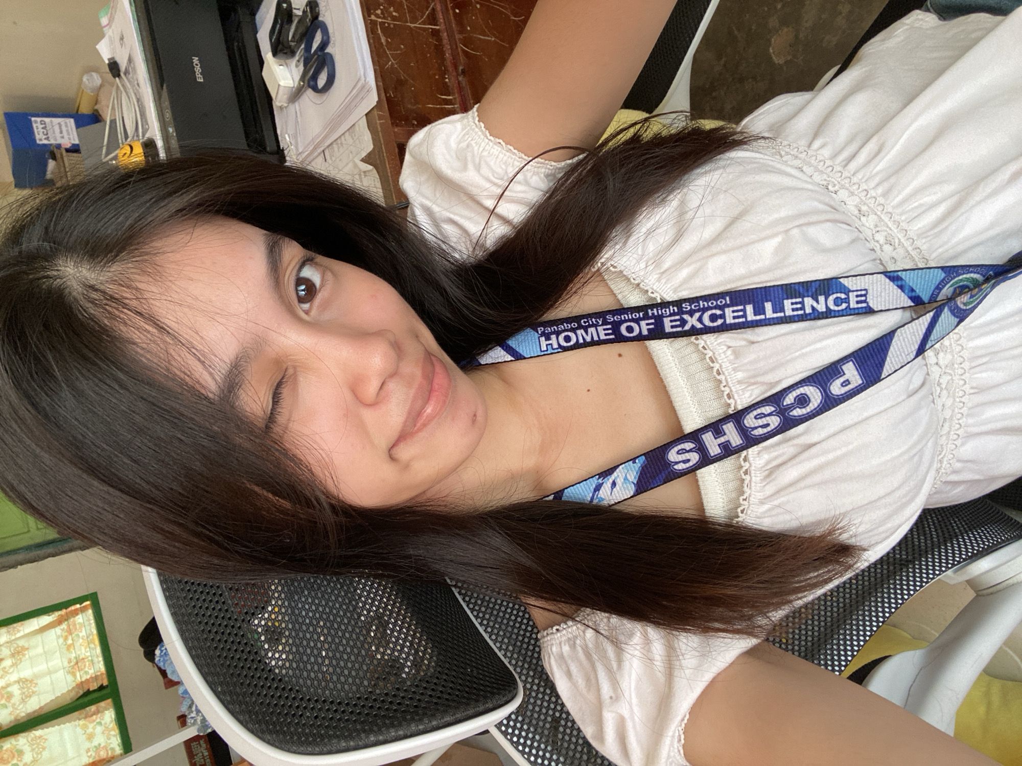 A young woman with long, dark hair is pictured winking at the camera in an indoor setting. She is wearing a white, off-the-shoulder top with ruffled details and a lanyard around her neck. The lanyard is blue and white, featuring the text "Panabo City Senior High School HOME OF EXCELLENCE". She is sitting in a black office chair, and behind her, a desk is visible with an Epson printer, papers, scissors, and a stapler. The background also shows a window with patterned curtains. The presence of "Panabo City Senior High School" on the lanyard suggests the location is Panabo City, Philippines.