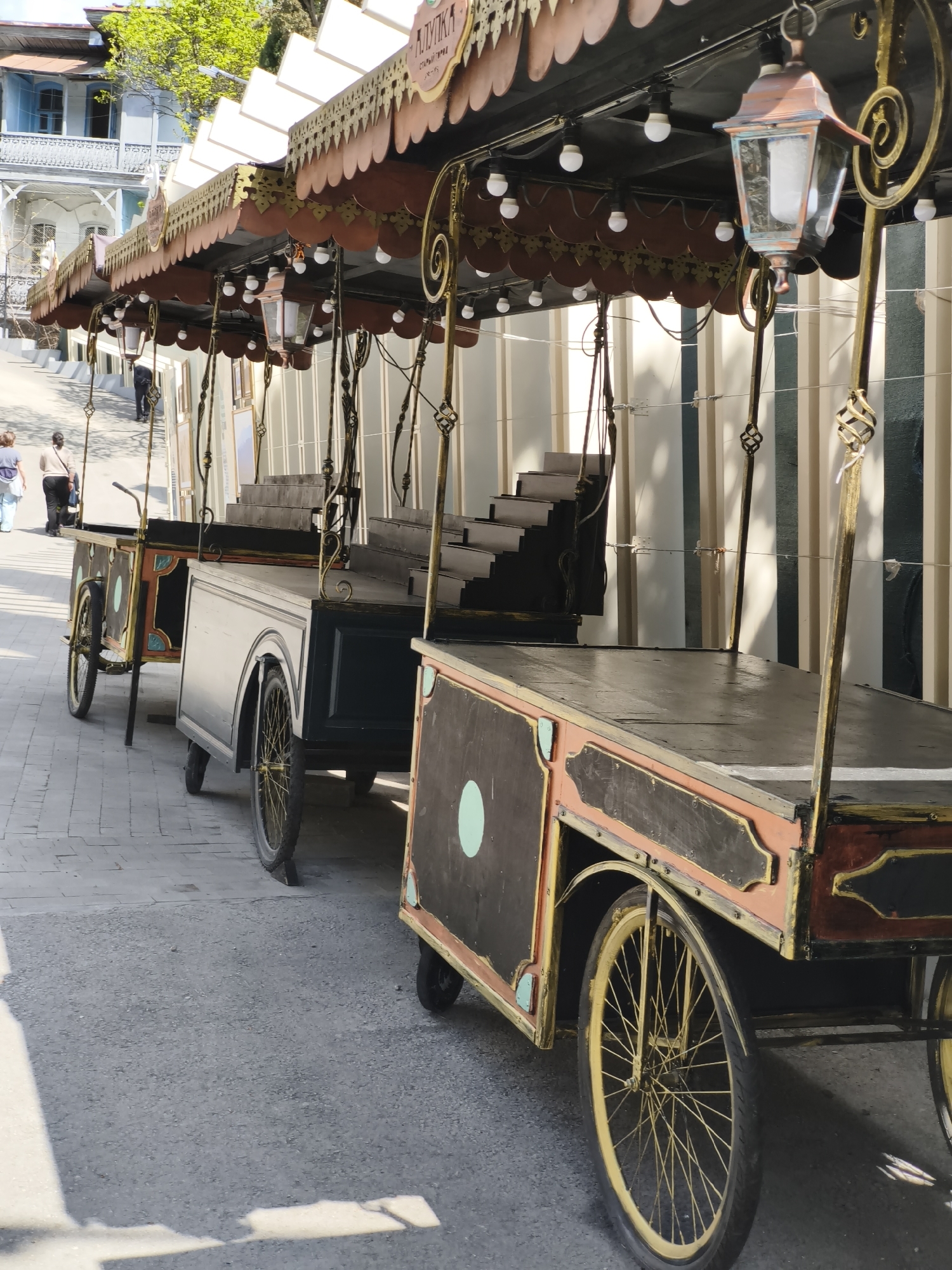 A street in Unknown, Unknown, has a row of ornamental carts parked along it. These carts are tricycle-style, with a large covered area above the main section of the cart, which features a sign in a foreign language. The sides of the carts are painted in a dark color with a light green circle on each side panel. Each cart is fitted with a vintage-style lantern hanging from the awning. Two women are walking on the pavement in the background. The carts are located alongside a building with a vertical striped exterior. The overall lighting is bright, suggesting it is daytime.