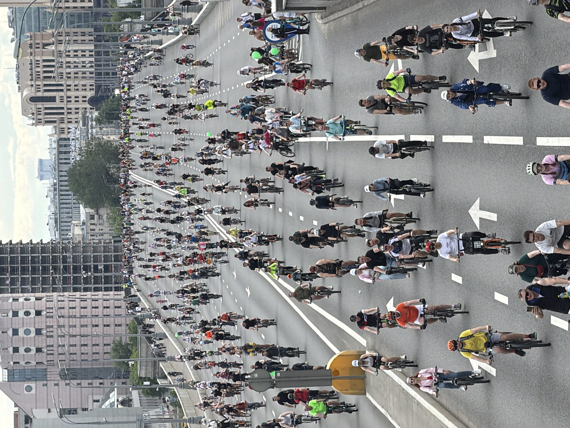 The image shows a large mass of cyclists participating in a bike parade, likely in the city of Moscow, Russia. They are riding on a wide roadway with multiple lanes, as indicated by the white lane markers and directional arrows. A mix of buildings and modern structures line the sides of the roadway. The cyclists are a diverse group of people, and they are riding in close proximity. The weather appears to be overcast or moderately sunny.